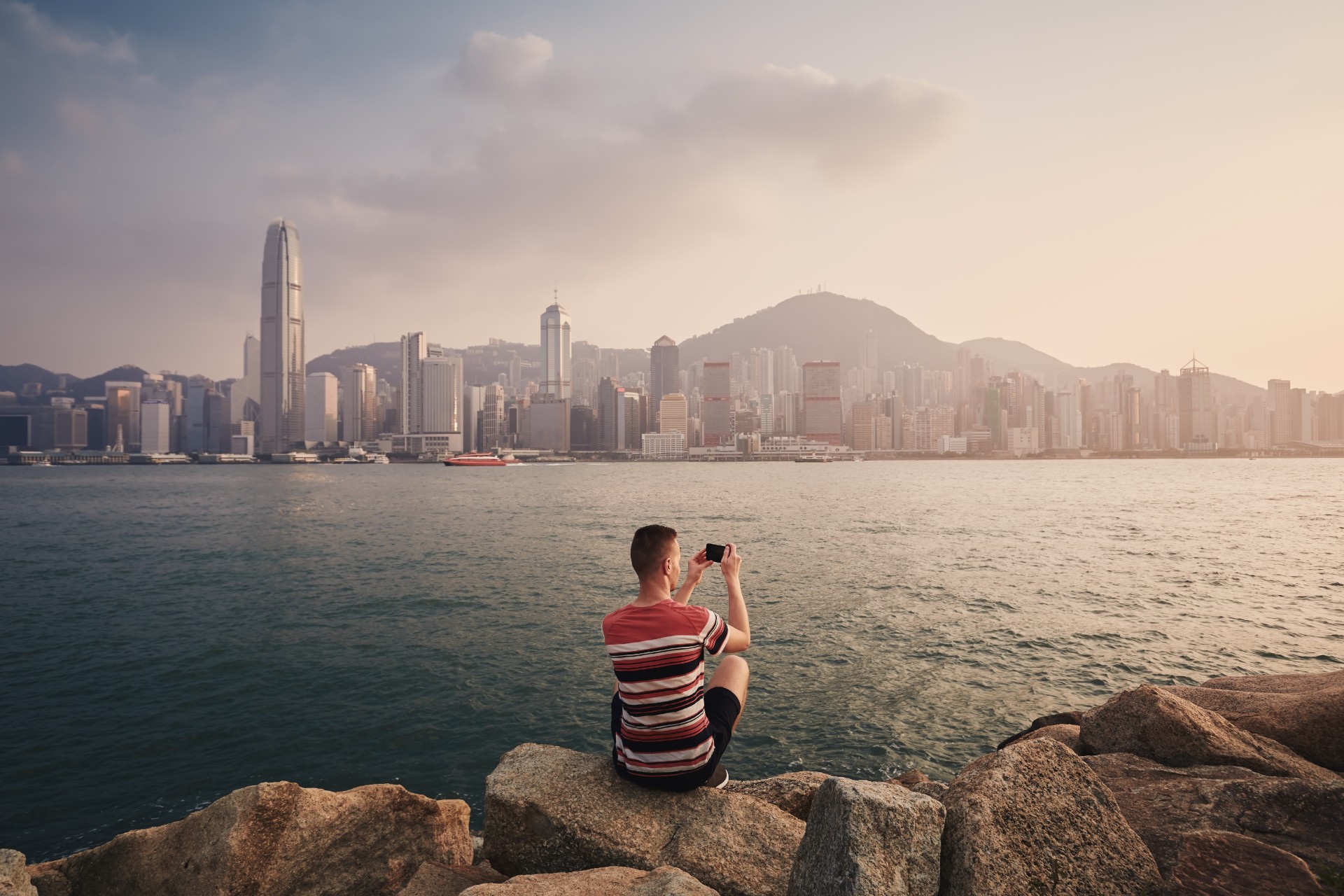 A person sitting on rocks by the water, taking a photo of the Hong Kong skyline at sunset.