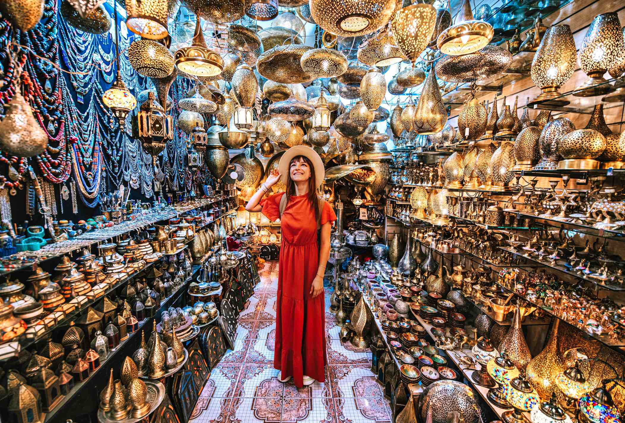 A woman in a red dress and hat stands in a shop filled with hanging lamps and decorative items.