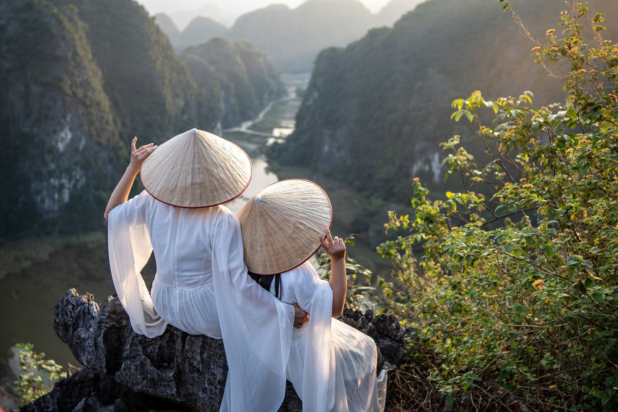 Two women in traditional conical hats sit on a rocky ledge, overlooking a scenic river and mountains at sunset.