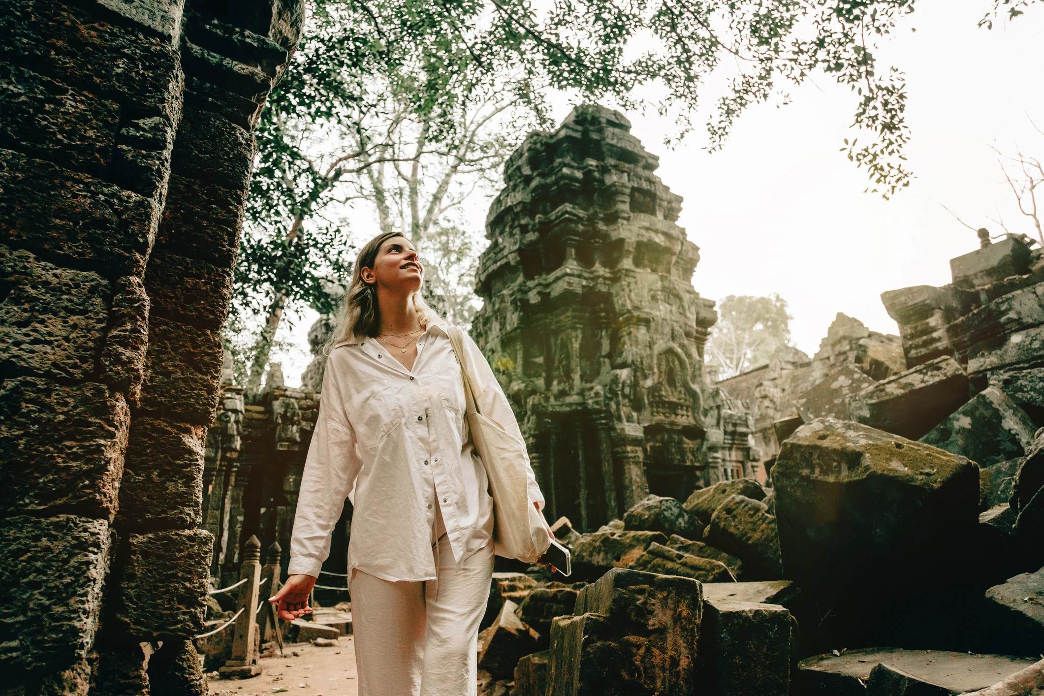 A woman in white clothing explores ancient ruins surrounded by overgrown trees and stone structures.