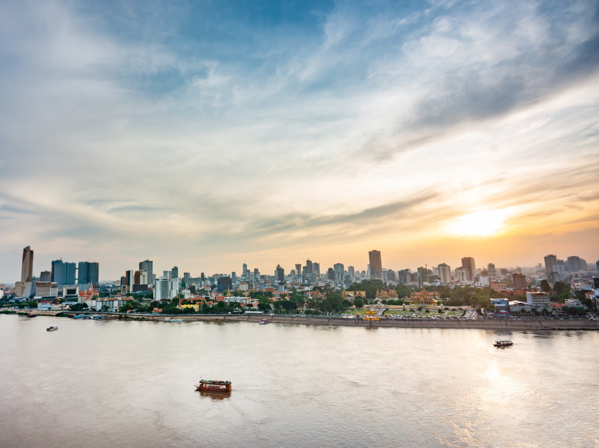 City skyline at sunset over a river, with clouds and boats visible on the water.