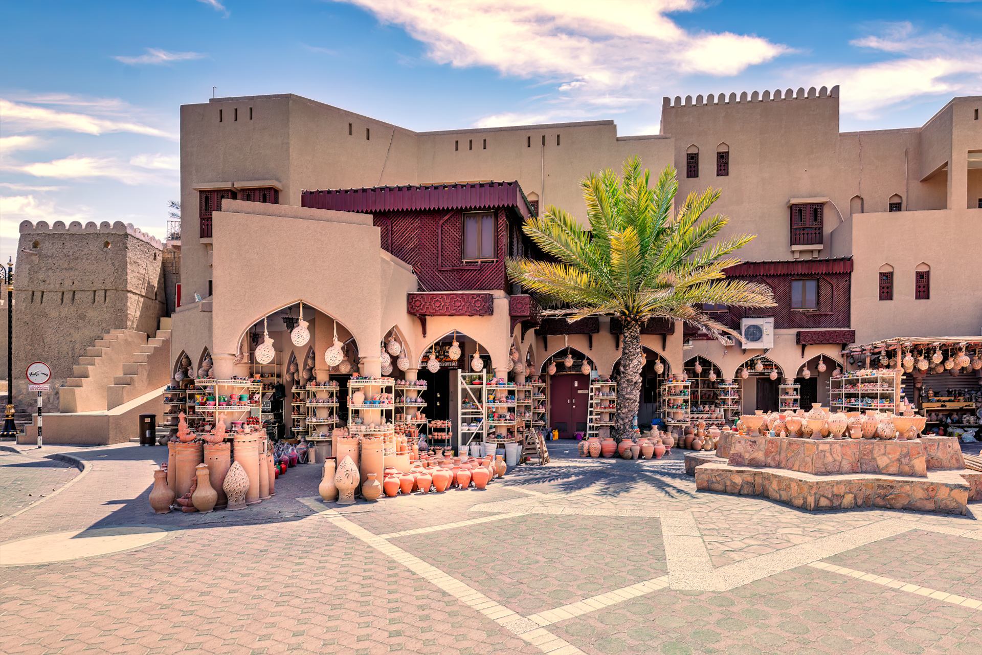 Market scene with clay pots for sale, palm trees, and traditional architecture under a blue sky.
