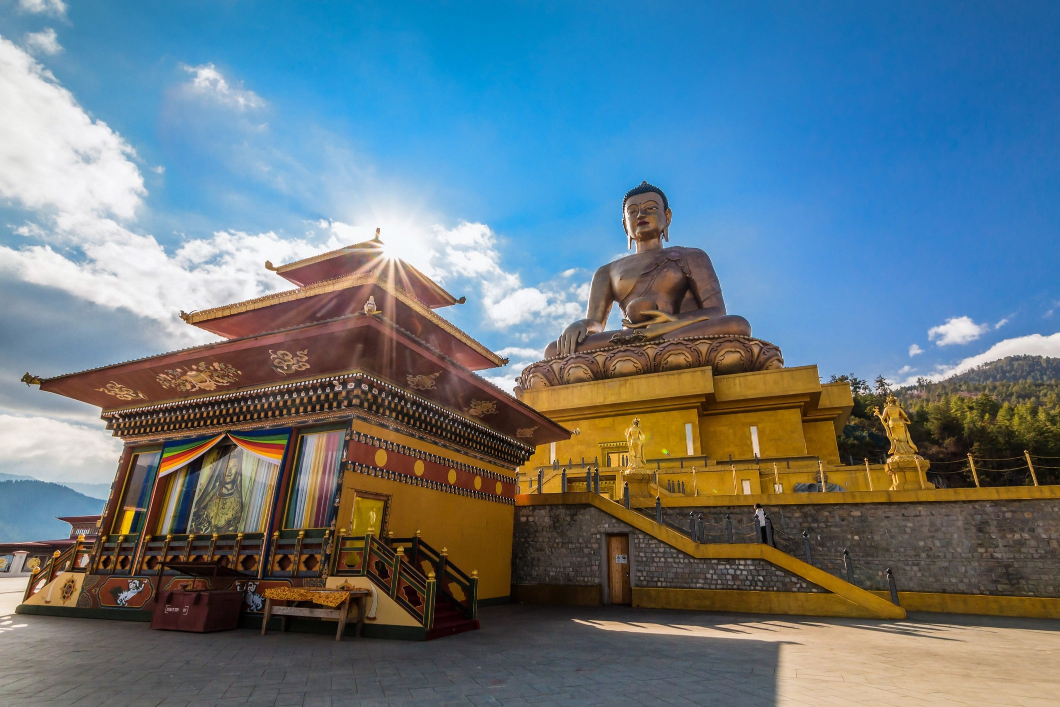 Golden Buddha statue beside a colorful temple under a bright sky, surrounded by mountains.