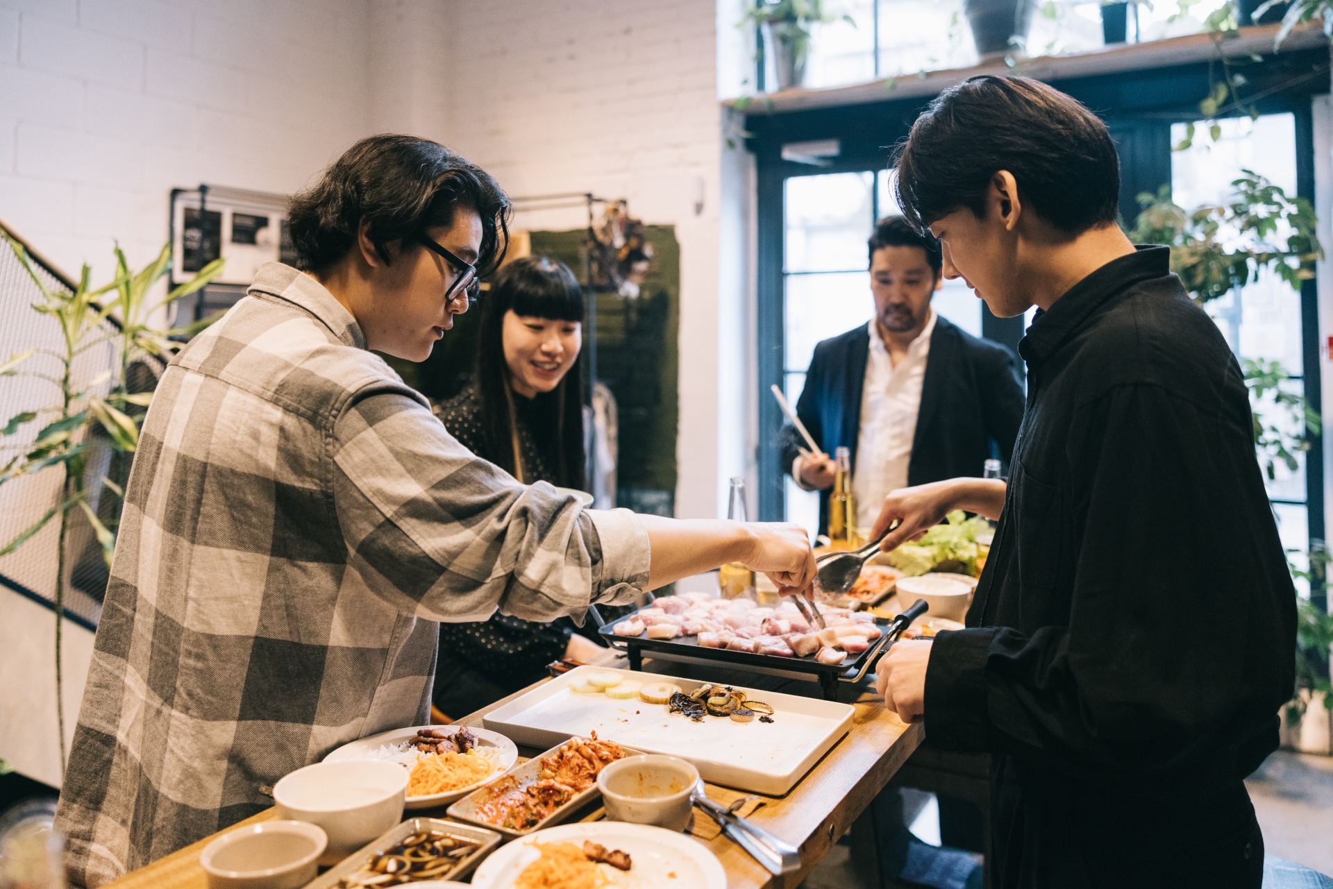 Group of friends enjoying a meal together, cooking at a table filled with various dishes and vibrant plants.