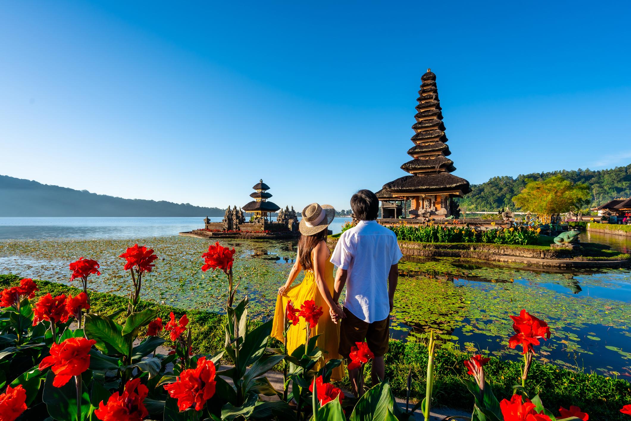 A couple holding hands by a lake, surrounded by vibrant flowers and a traditional pagoda in the background under a clear blue sky.