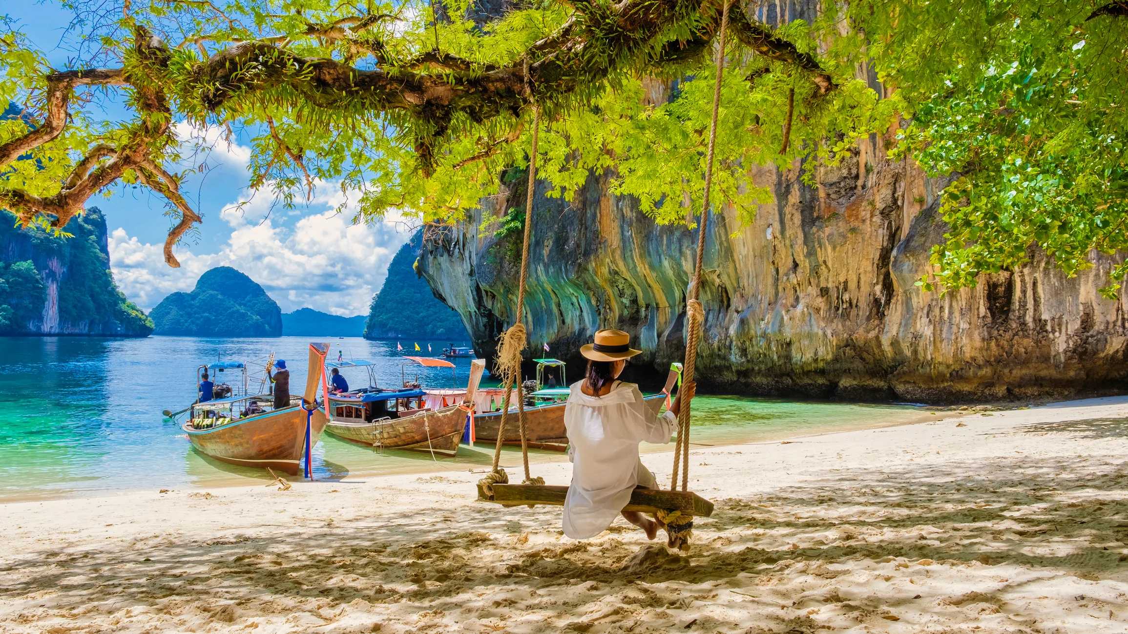 A person on a swing overlooking a tropical beach with boats and limestone cliffs in the background under a sunny sky.