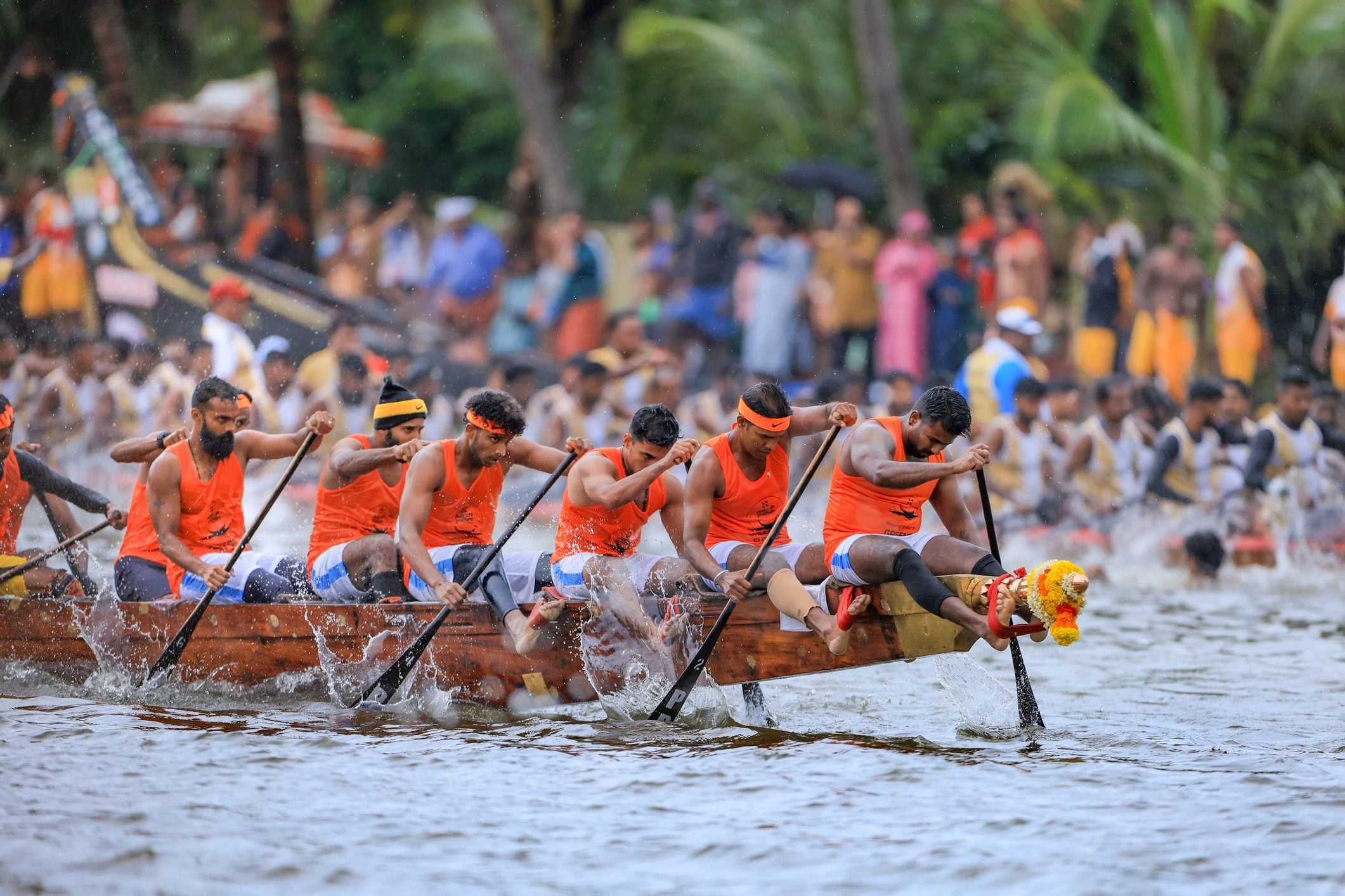 A team of rowers in orange shirts paddles a dragon boat, competing in a race with a large crowd watching.