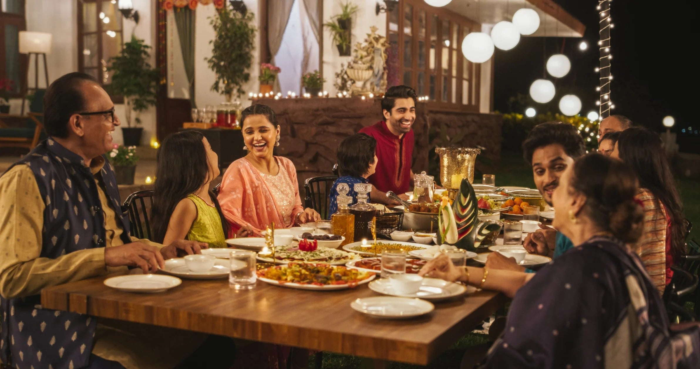 A festive outdoor dinner with several people enjoying food, laughing, and celebrating under soft lighting.