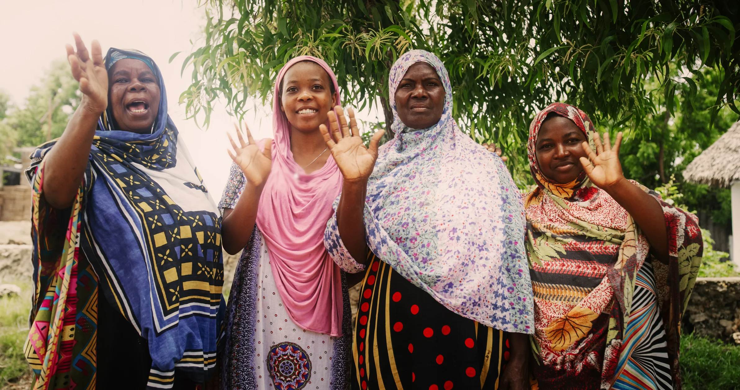 Four women in colorful dresses wave and smile, standing together under a tree in a vibrant outdoor setting.