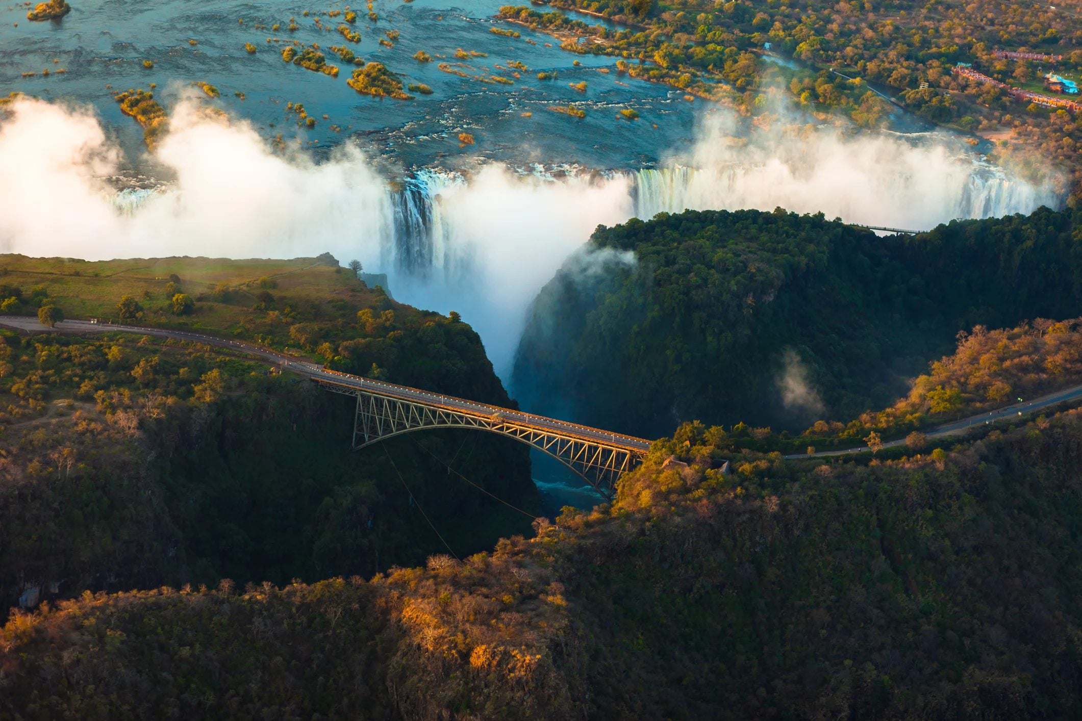 Aerial view of a waterfall with mist, featuring a bridge spanning a canyon surrounded by lush greenery.