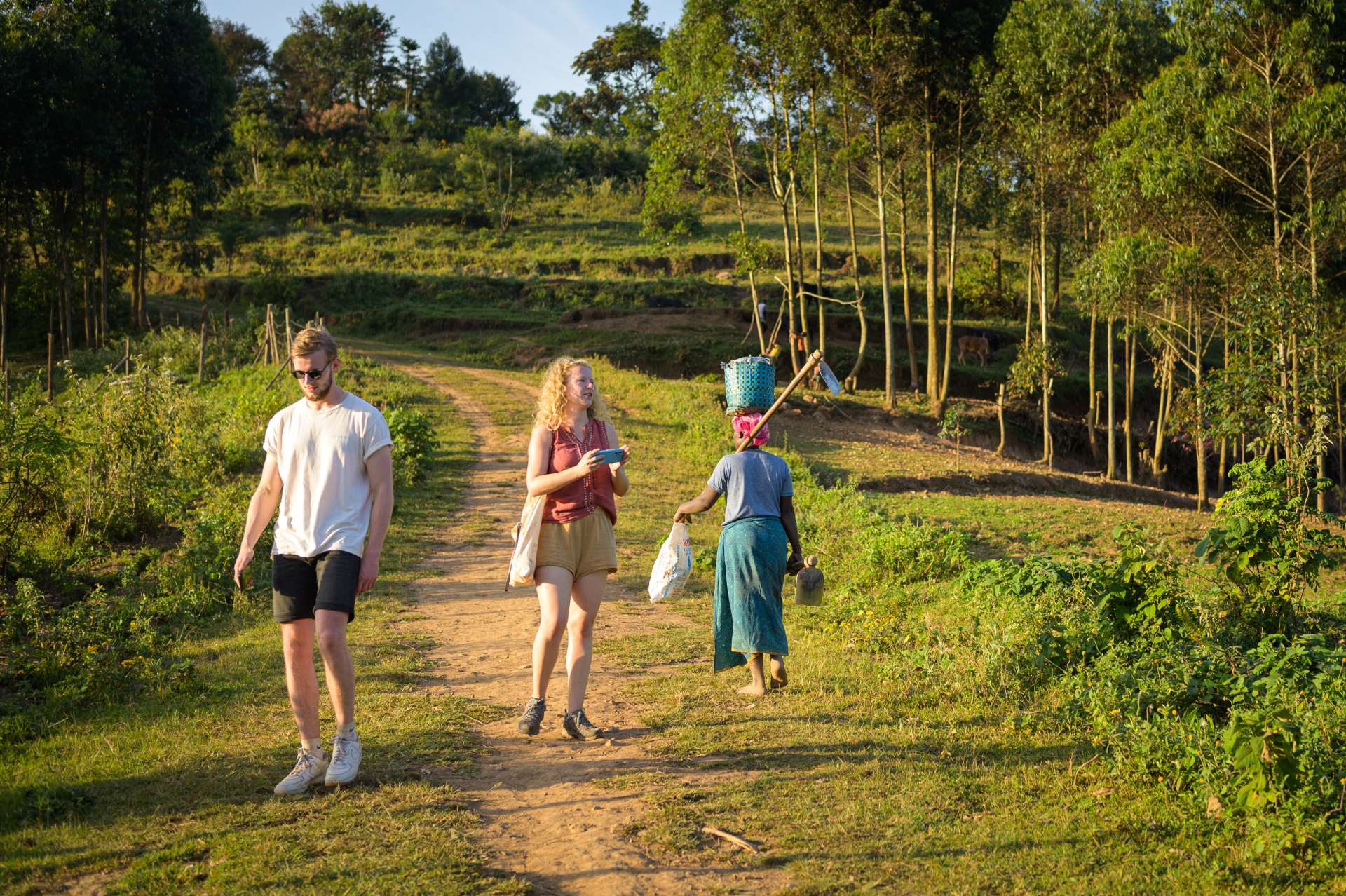 Two people walk on a dirt path in a rural setting, while a woman carries items in a basket nearby. Lush greenery surrounds them.