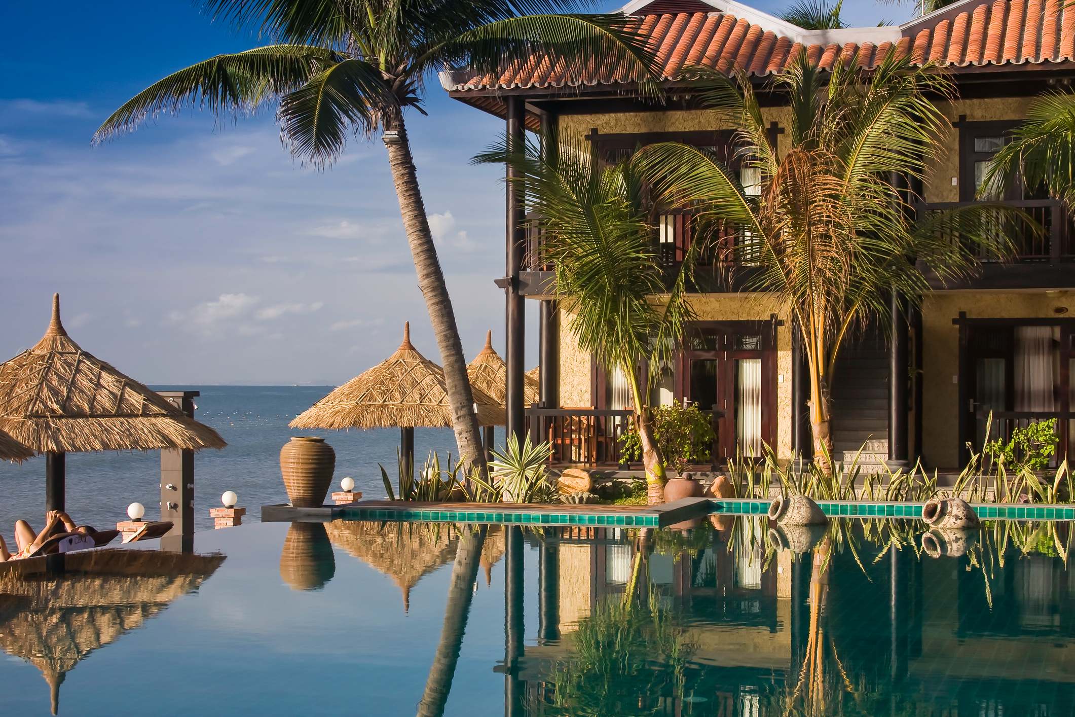 Tropical beachfront villa with palm trees, a pool, and thatched umbrellas overlooking the ocean at sunset.
