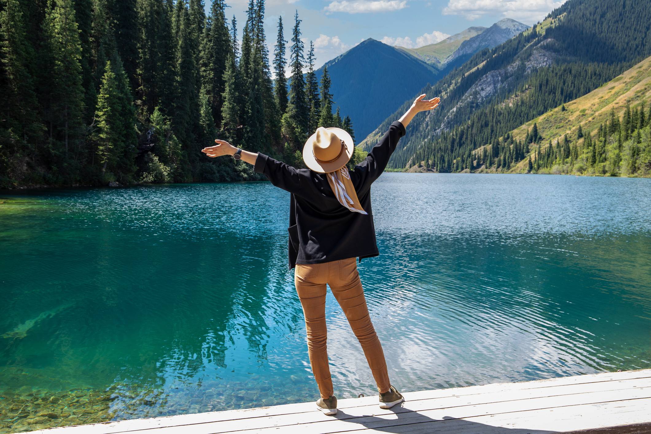 Person in a hat standing by a serene blue lake, arms outstretched, surrounded by trees and mountains under a clear sky.