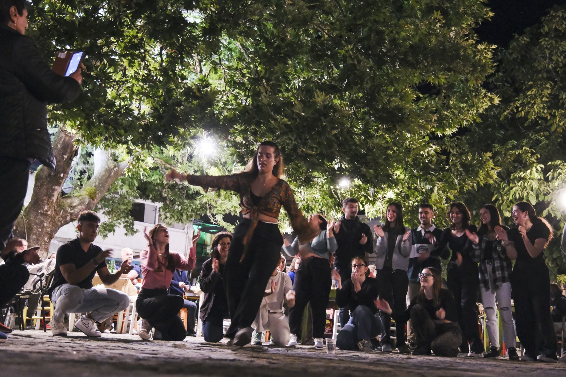 A lively outdoor scene with a dancer performing, surrounded by an engaged audience, under bright lights and trees.