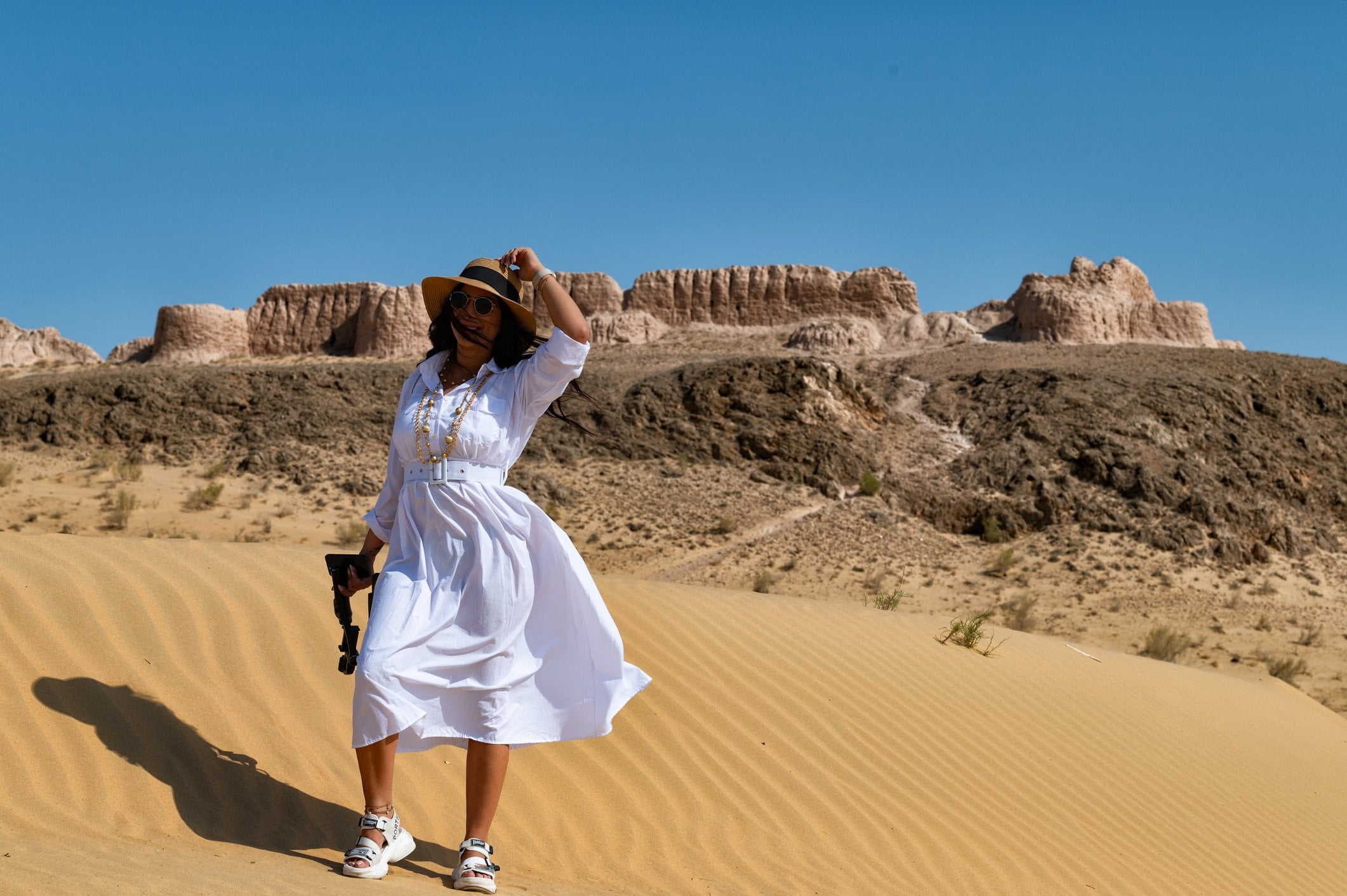 A person in a white dress and hat stands on sand dunes with desert cliffs in the background, holding a camera.