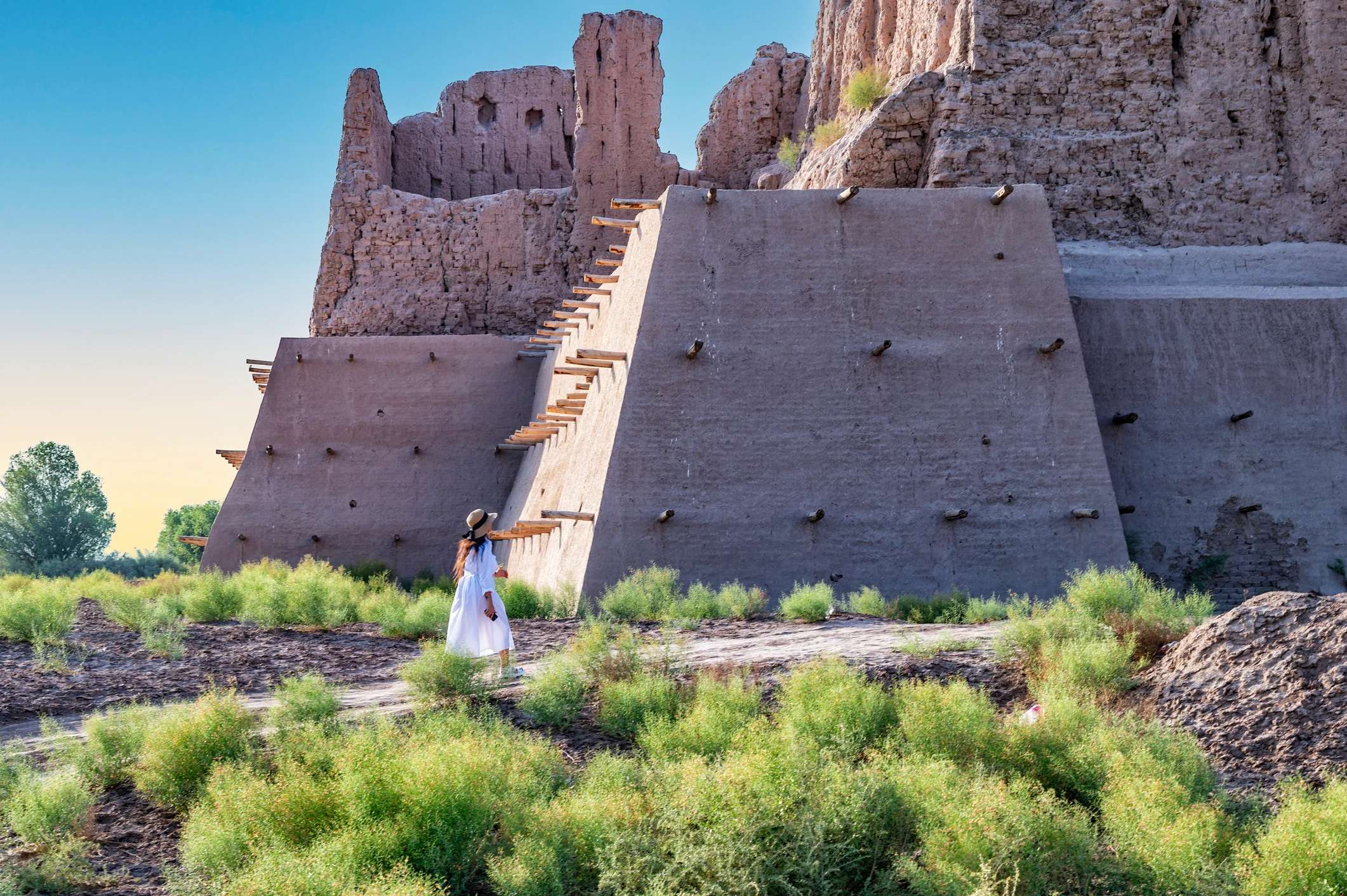 A person in a white dress stands near a large, ancient structure surrounded by green vegetation under a clear sky.