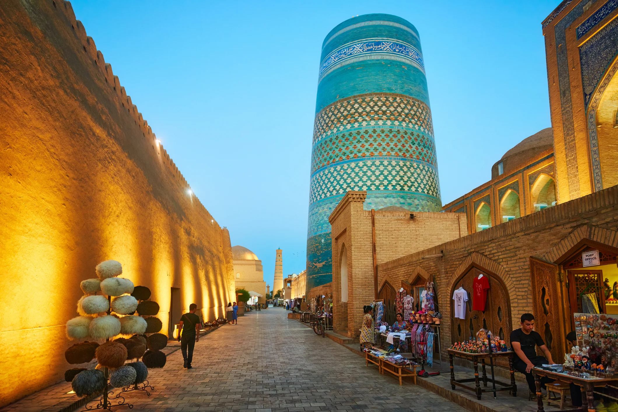 Evening scene in Khiva, Uzbekistan, showcasing a historic street lined with shops and a prominent blue minaret.
