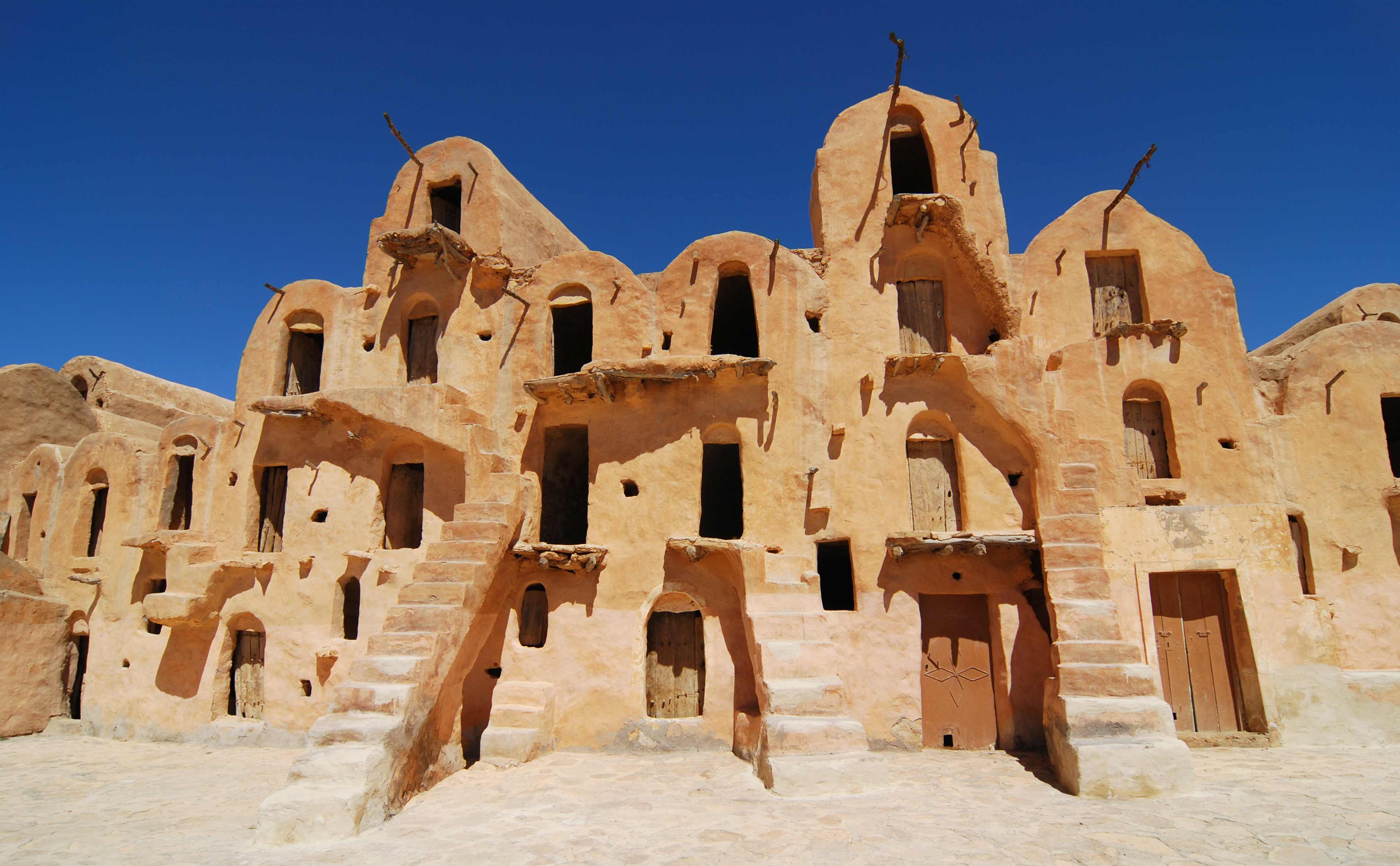 Ancient desert architecture featuring multi-level, ochre-colored buildings with stairs and numerous windows under a blue sky.
