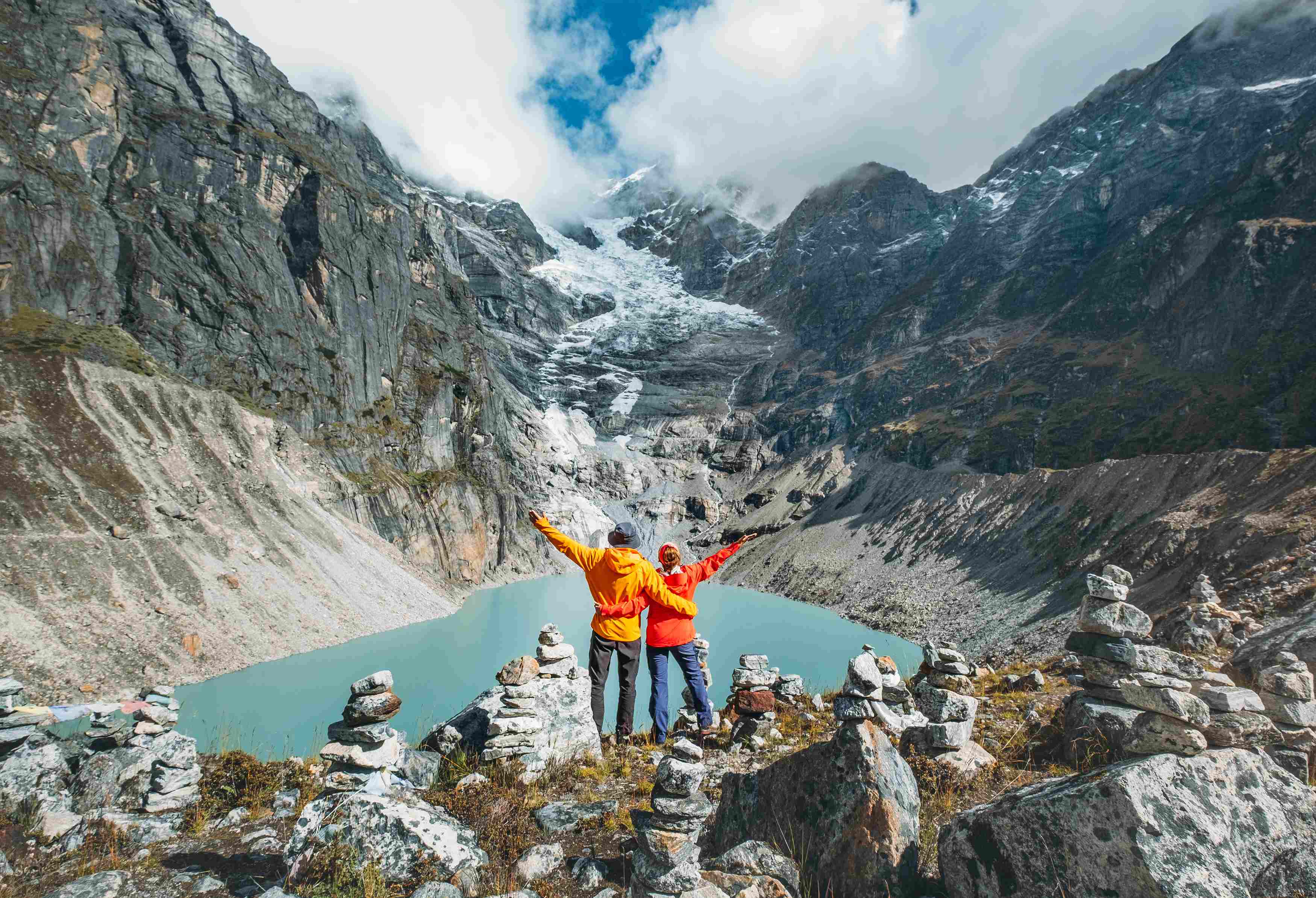 Two hikers in bright orange jackets stand atop a rocky overlook, celebrating the stunning mountain landscape and glacial lake below.
