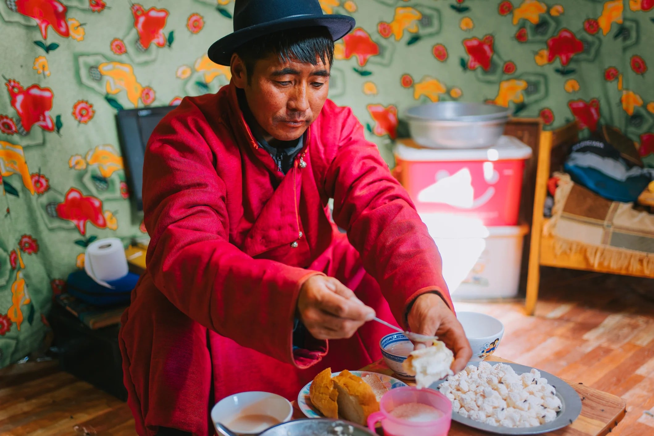 A man in a red robe serves food from a plate while sitting indoors, surrounded by colorful patterned walls.