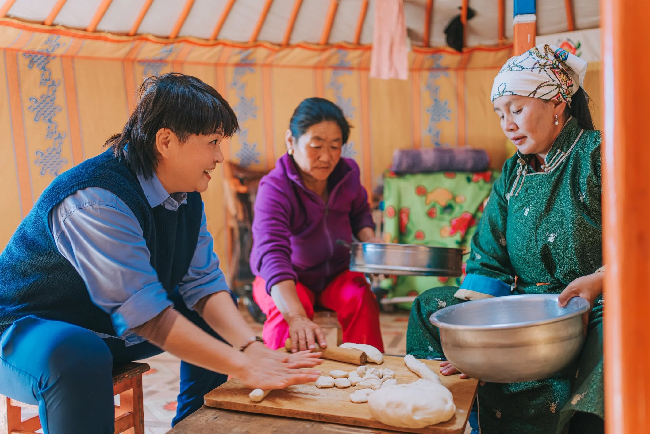 Three people prepare food inside a traditional yurt, with one person rolling dough and the others assisting at a table.