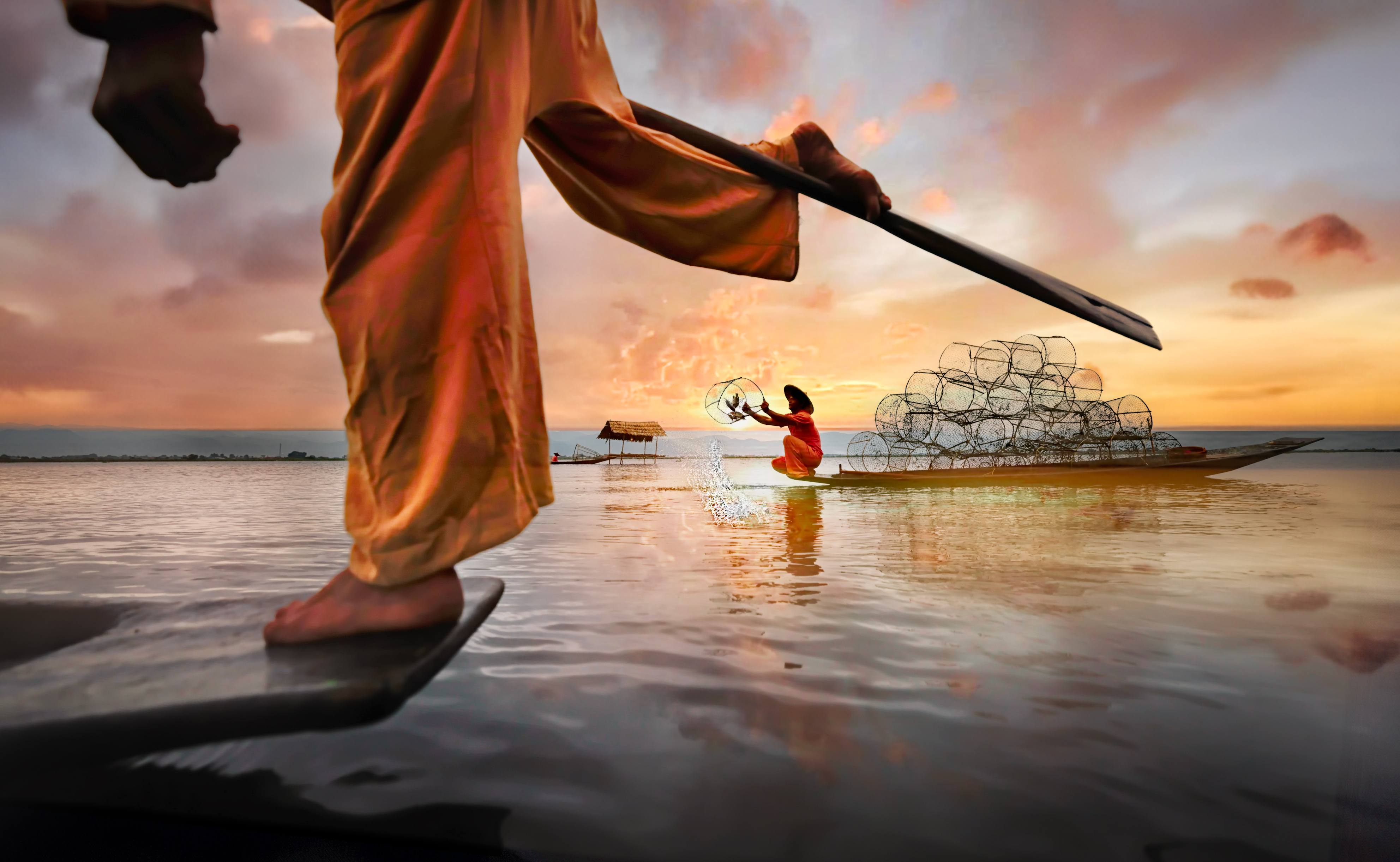A fisherman in traditional dress uses a spear, while a woman in a boat casts nets at sunset over calm water.