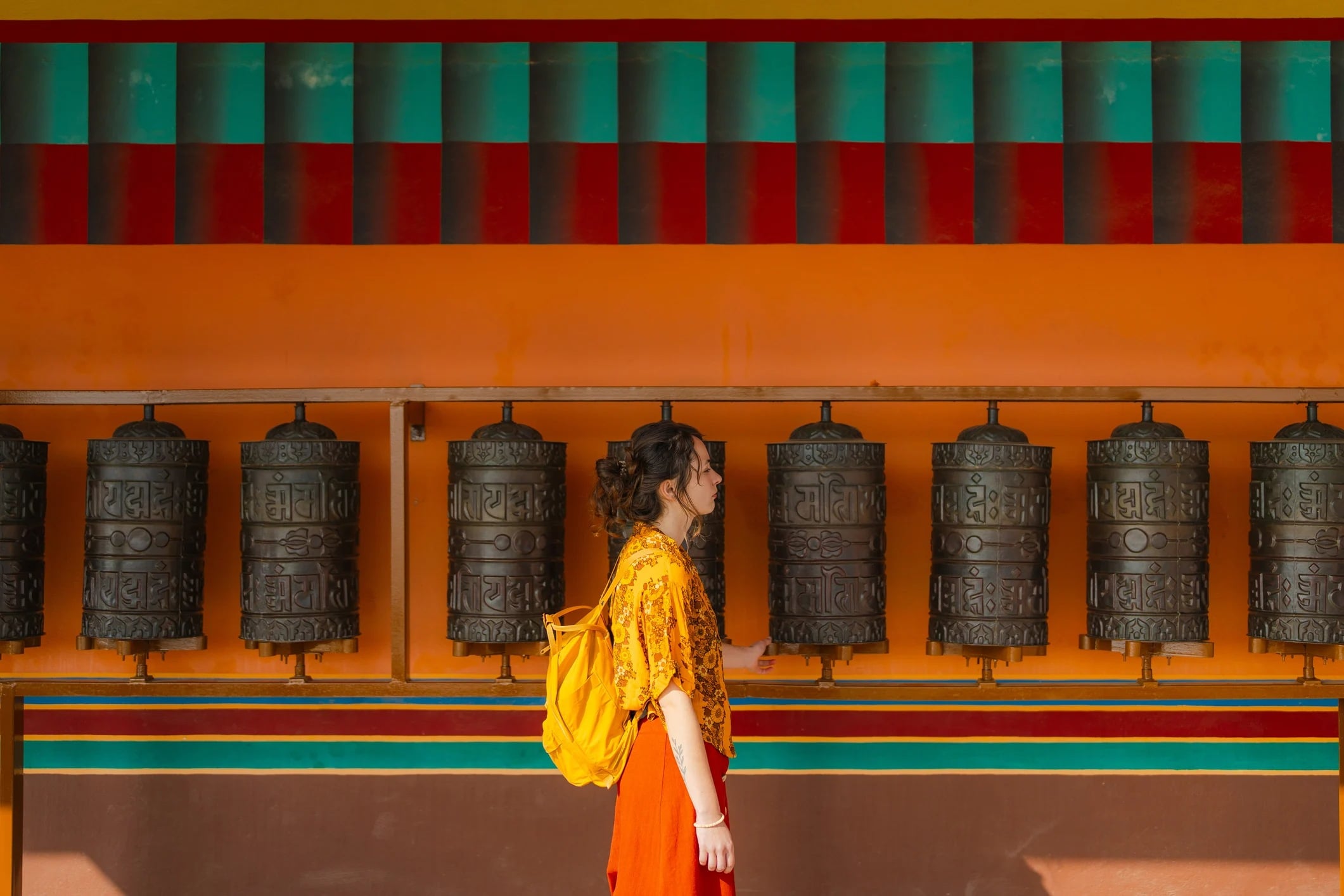 A woman in a yellow shirt and orange skirt walks past a row of prayer wheels against a vibrant orange and turquoise wall.