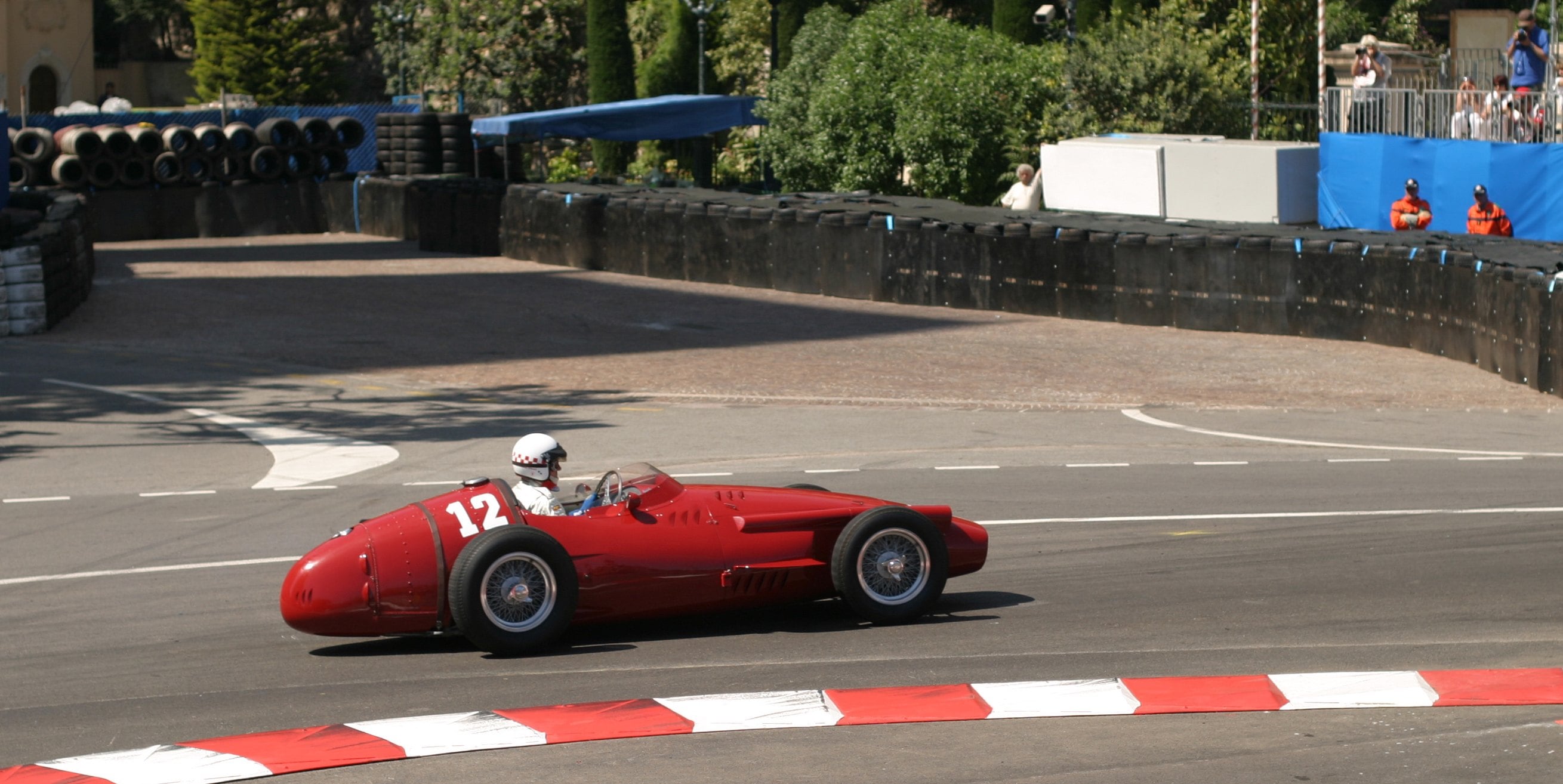 A vintage red race car, number 12, speeds around a bend on a racetrack with spectators in the background.