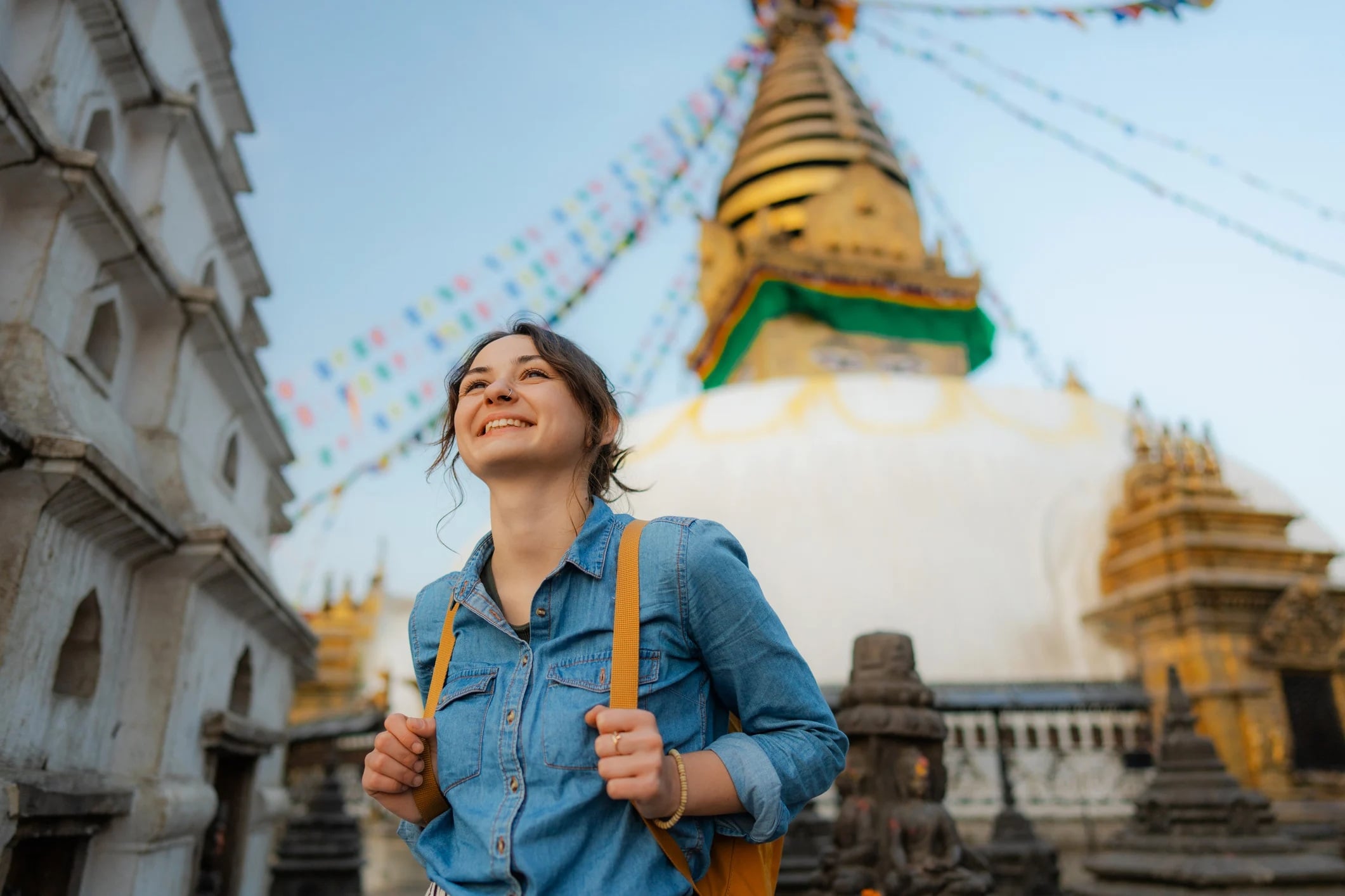 Smiling traveler in a denim jacket stands at a temple with colorful prayer flags and a stupa in the background.