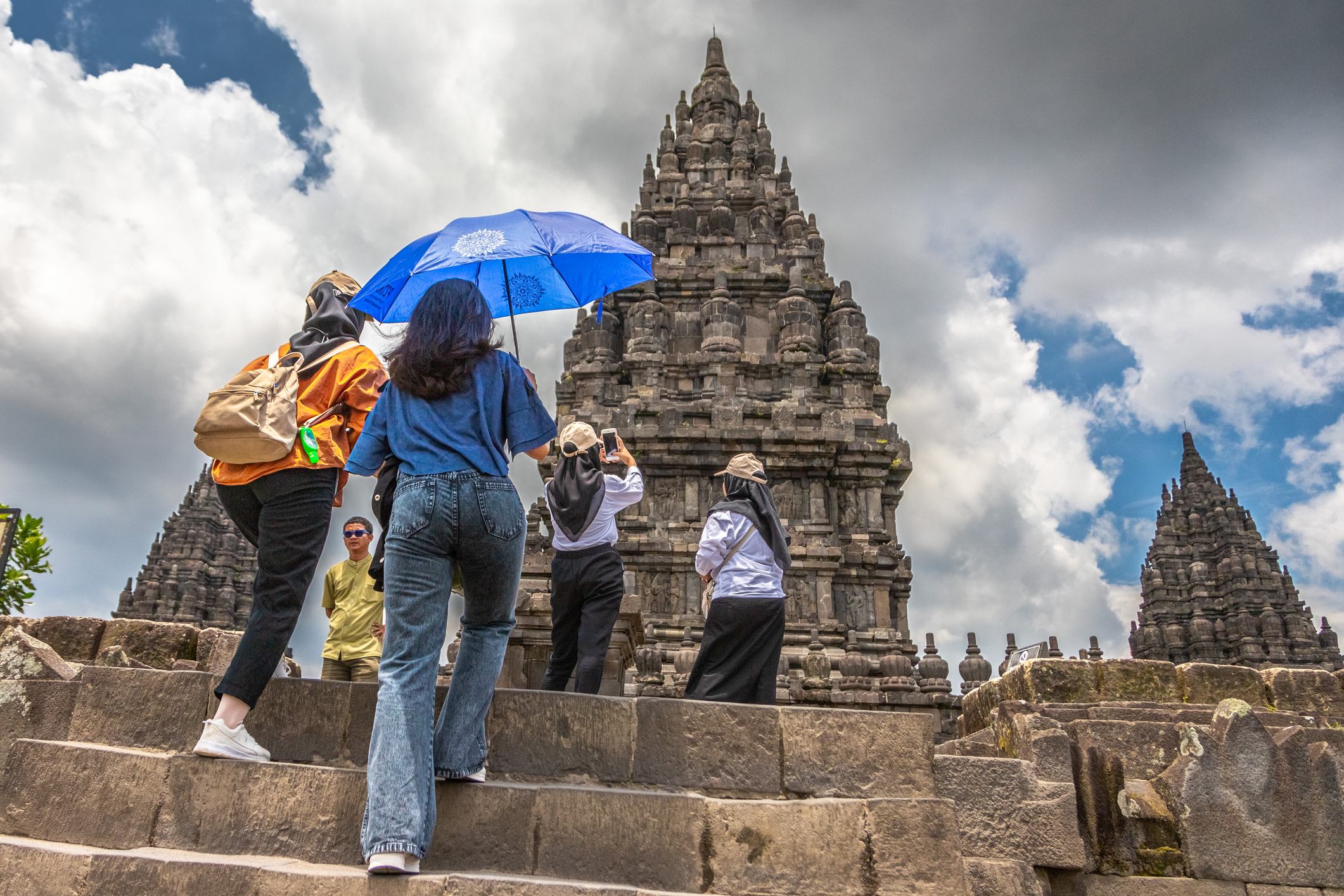 Visitors ascend stone steps toward ancient temples under a partly cloudy sky, some using umbrellas for shade.