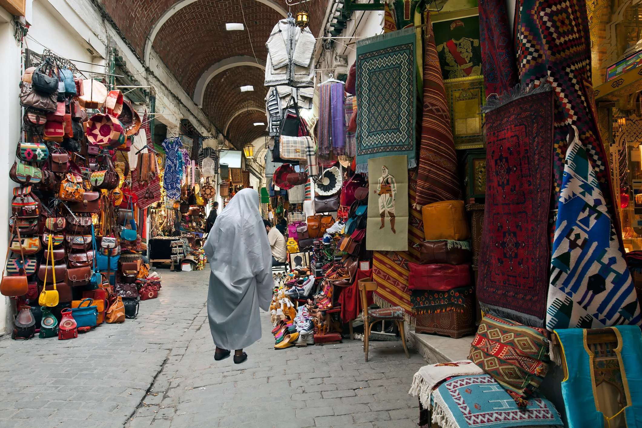 A bustling market with vibrant textiles, bags, and crafts, featuring a person in a gray cloak walking through the narrow path.