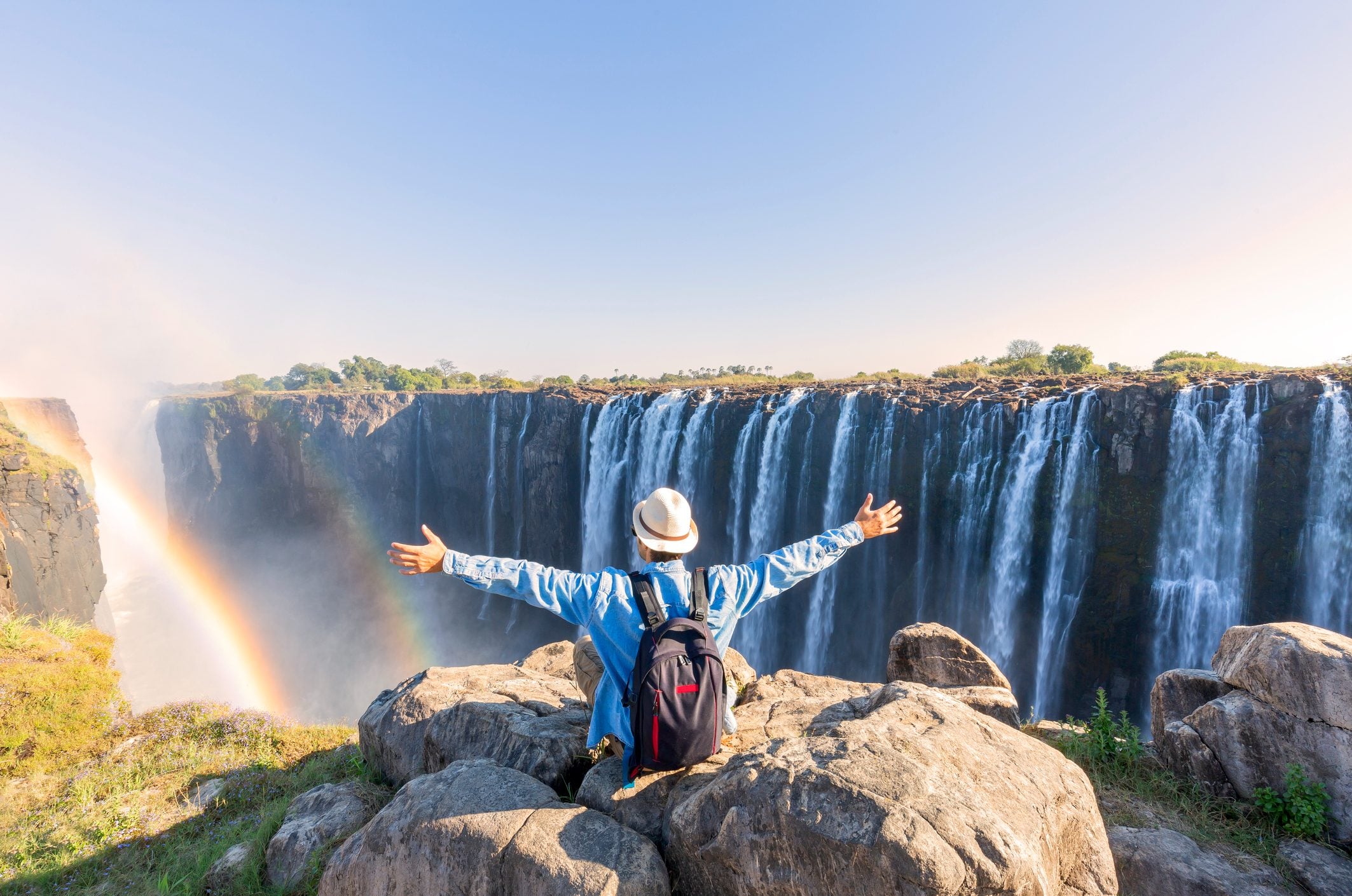 Person with a backpack and hat standing on rocks, arms outstretched, overlooking a majestic waterfall with a rainbow.