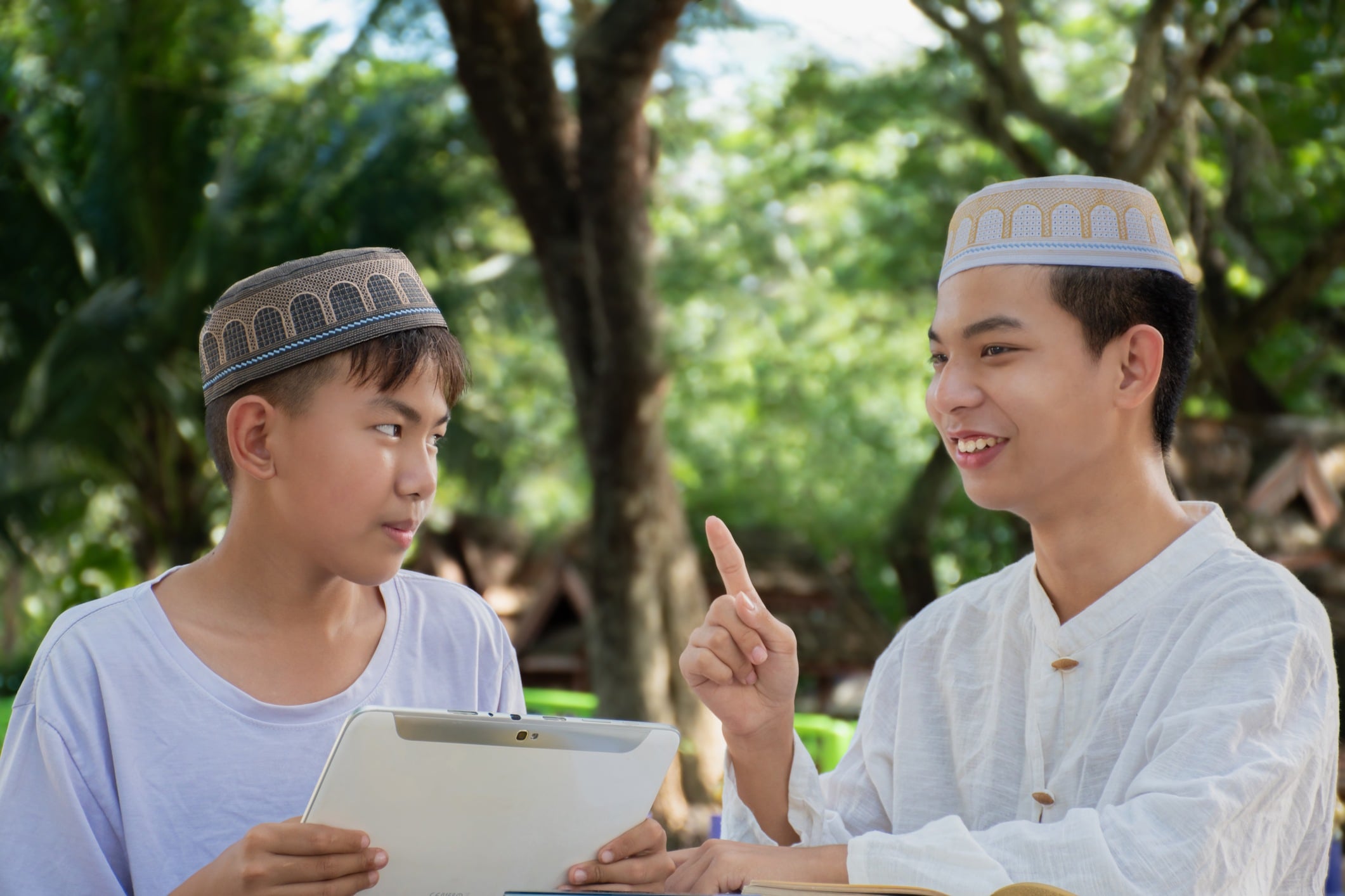 Two young men wearing traditional hats engage in conversation outdoors, one holding a tablet and listening intently.