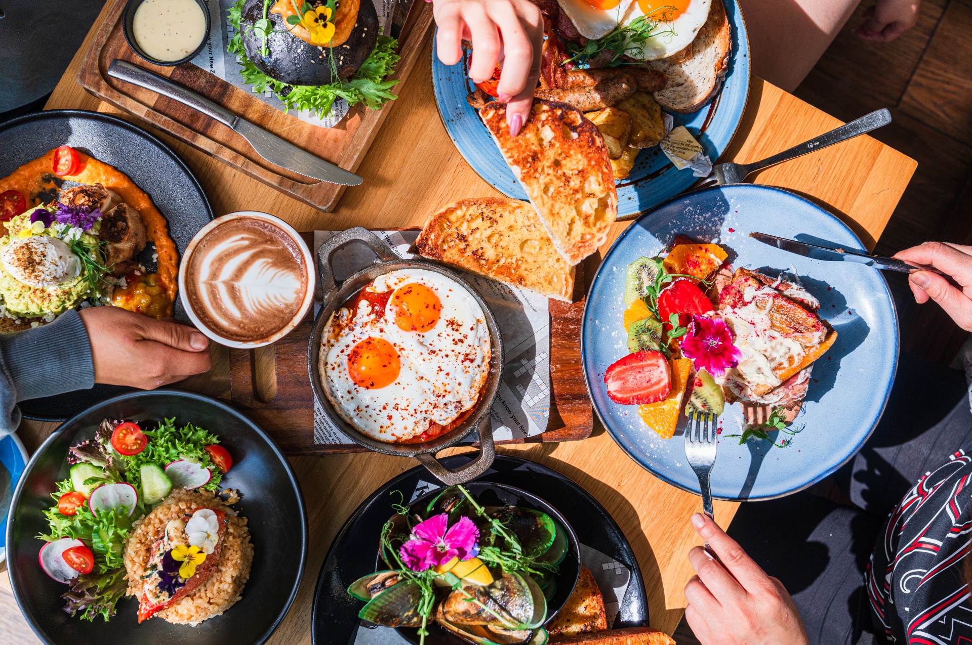 A top view of a diverse brunch spread with dishes, drinks, and hands sharing food around a wooden table.