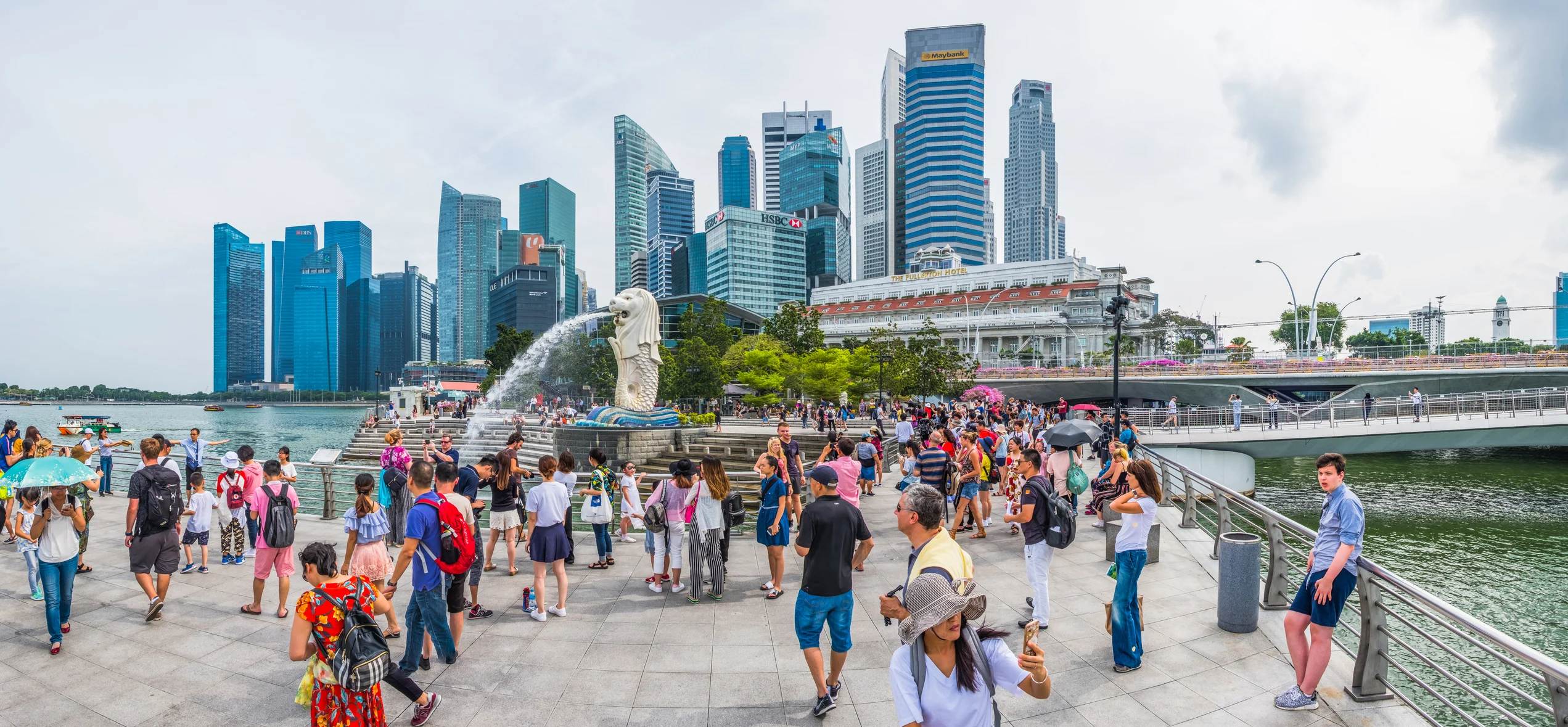 Crowd gathered by the Merlion statue with Singapore's skyline in the background, featuring modern skyscrapers.