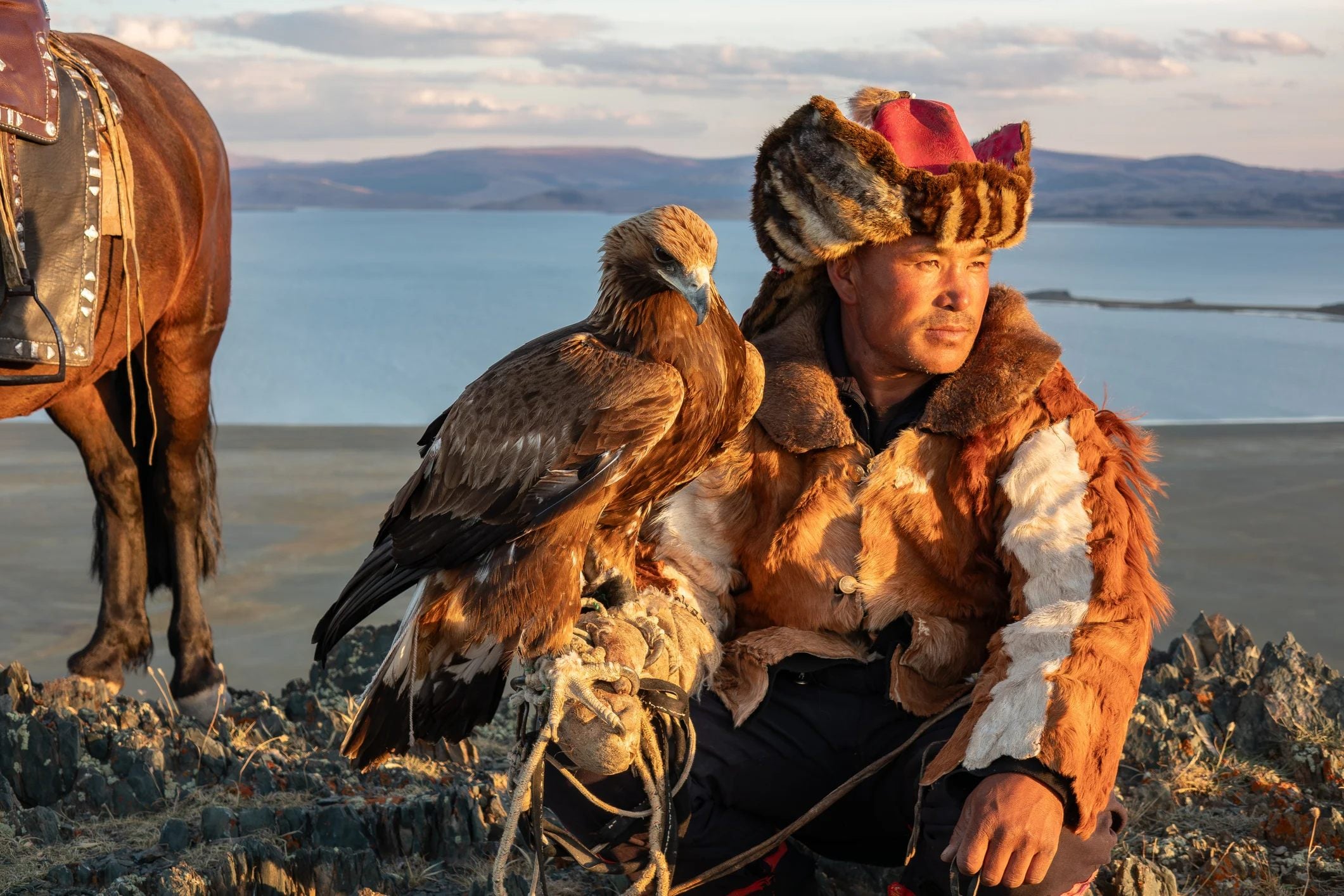 A hunter in traditional attire sits with a golden eagle, overlooking a scenic landscape with a horse nearby.