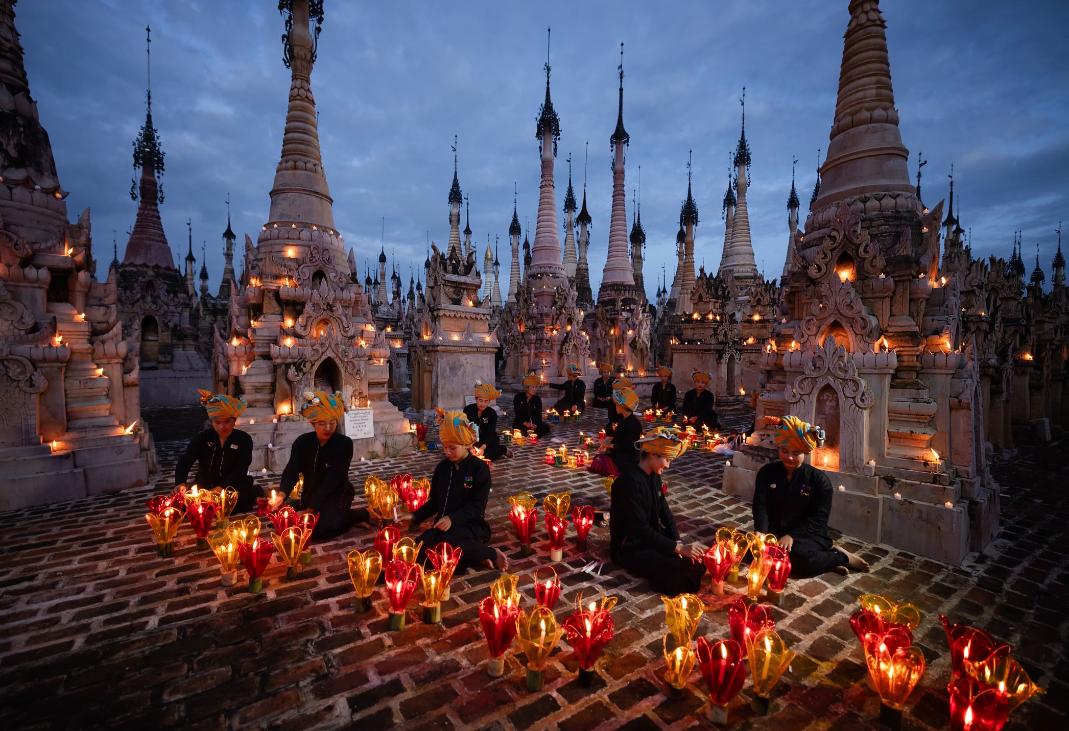 A serene twilight scene with people seated among illuminated pagodas and glowing candles, creating a peaceful atmosphere.