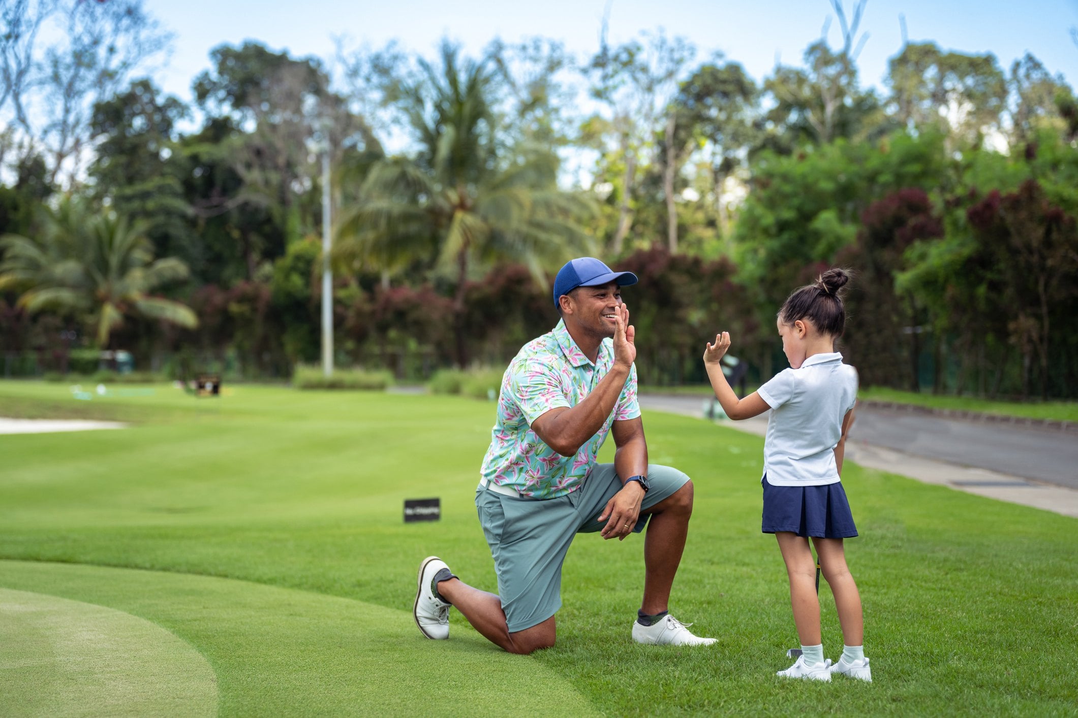 A man kneels on a golf course while a girl, wearing a golf outfit, playfully gestures with her hand.