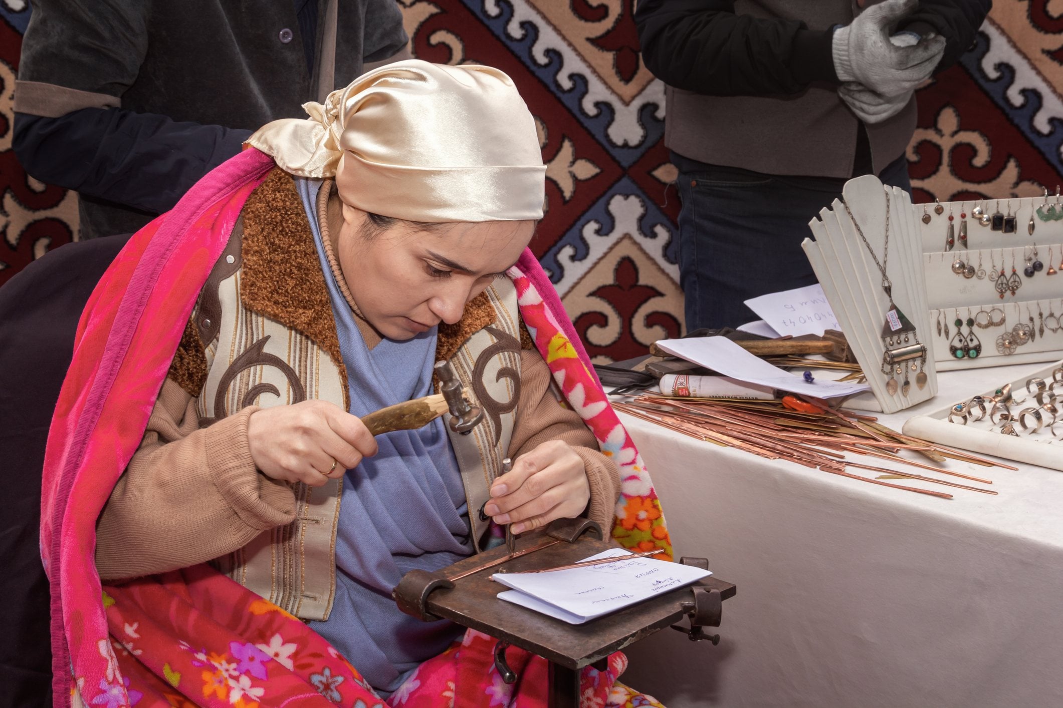 A woman in traditional attire works on a craft at a table with various tools and decorative items in the background.