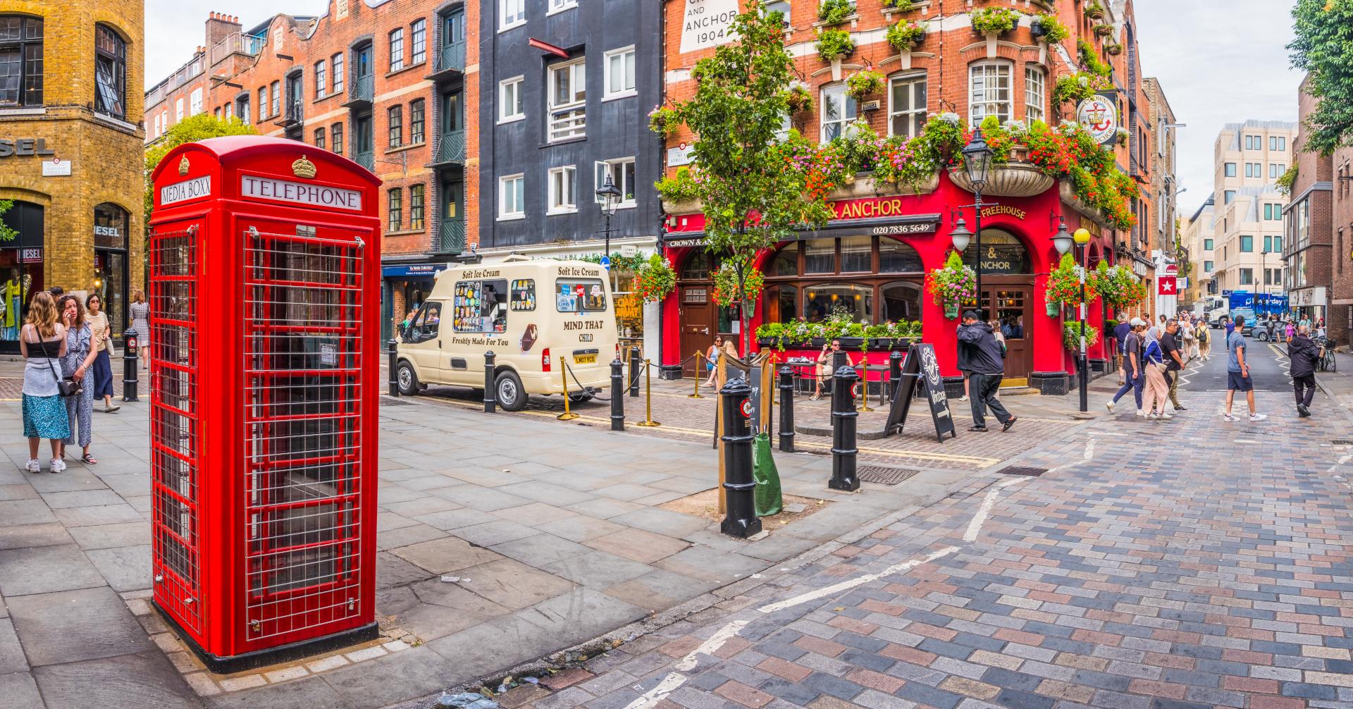 A vibrant street scene featuring a red telephone box, a pub adorned with flowers, and people enjoying the urban atmosphere.
