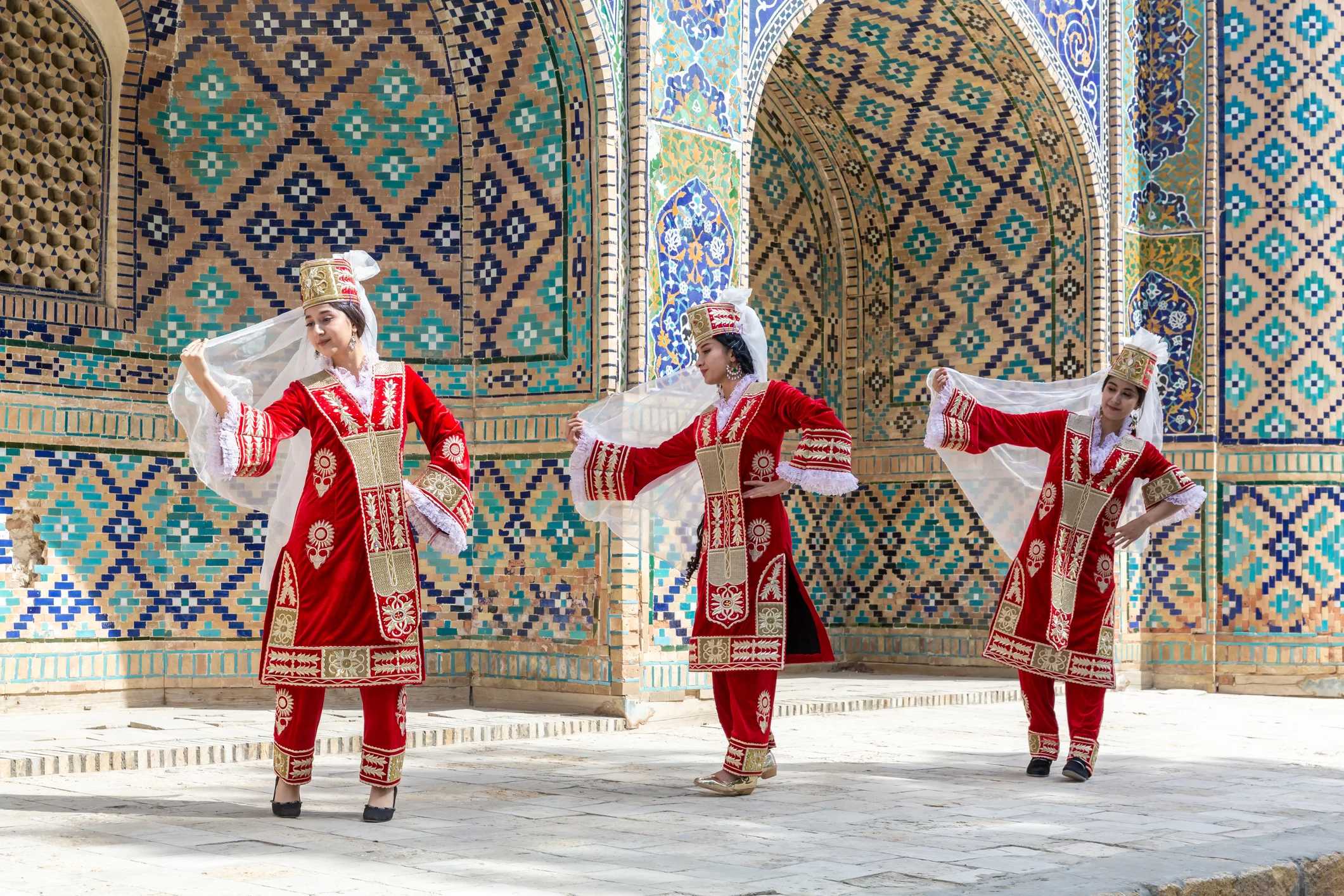 Three dancers in traditional red costumes perform against a colorful, tiled backdrop. They hold flowing white veils.