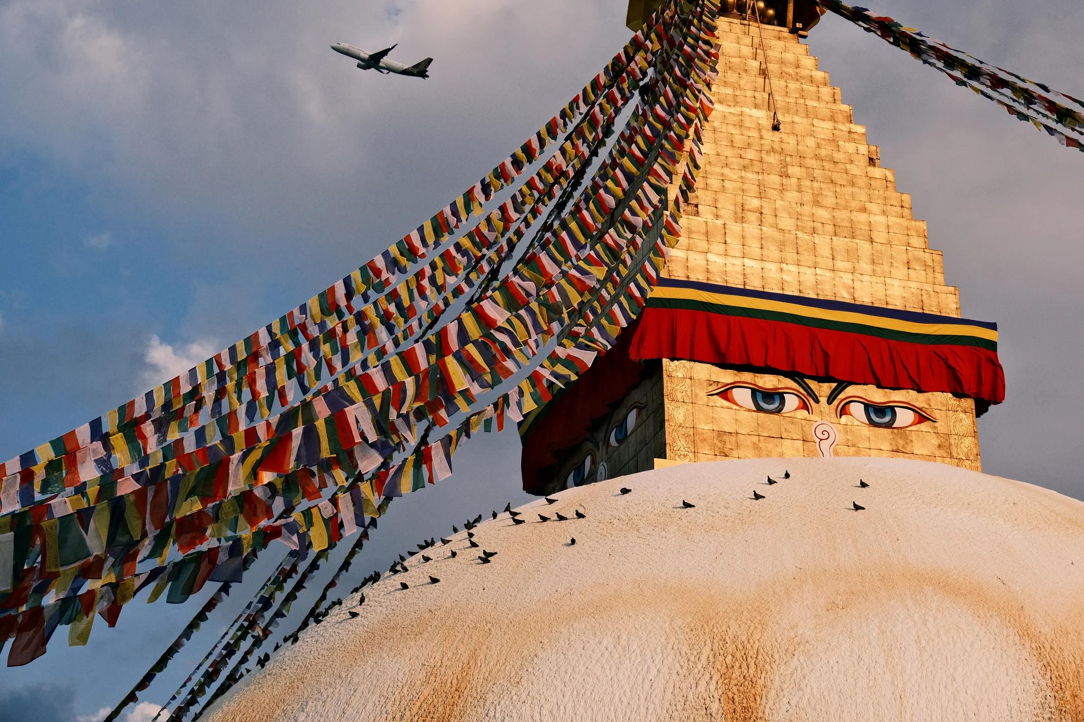 Boudhanath Stupa, adorned with colorful prayer flags, stands under a cloudy sky, with an airplane flying overhead.