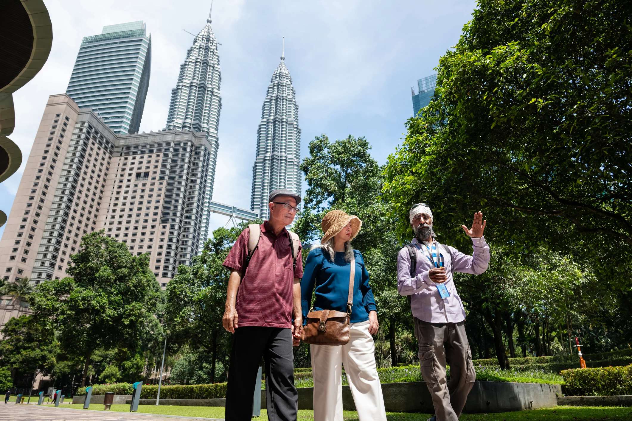 Three people walk together in a park with tall buildings visible in the background under a clear blue sky.