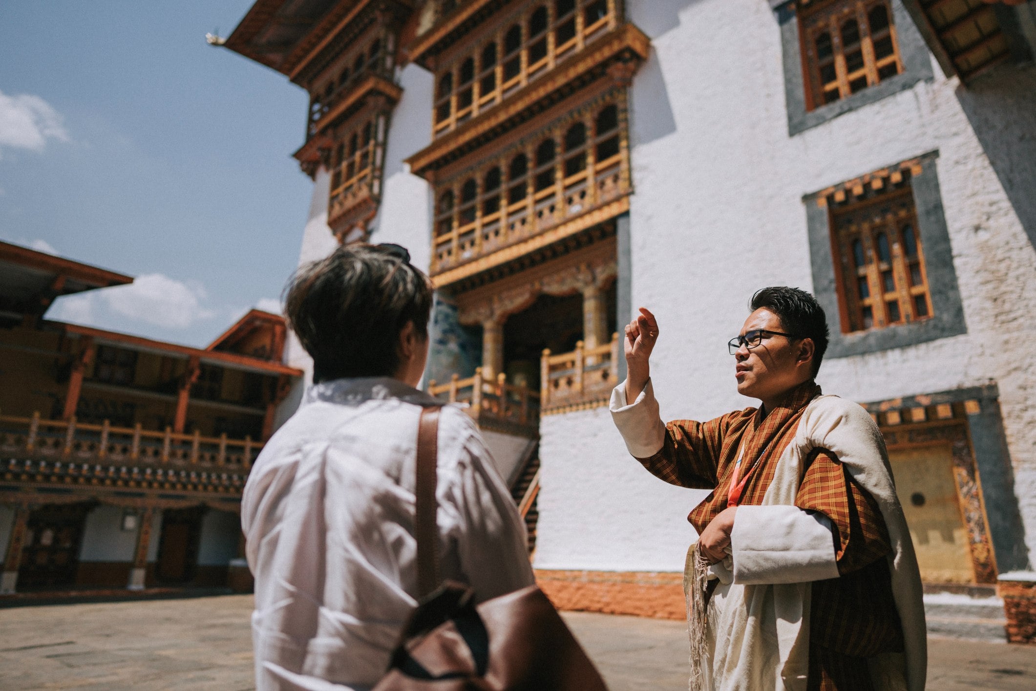 A tour guide gestures while speaking to a visitor outside a traditional Bhutanese building on a sunny day.