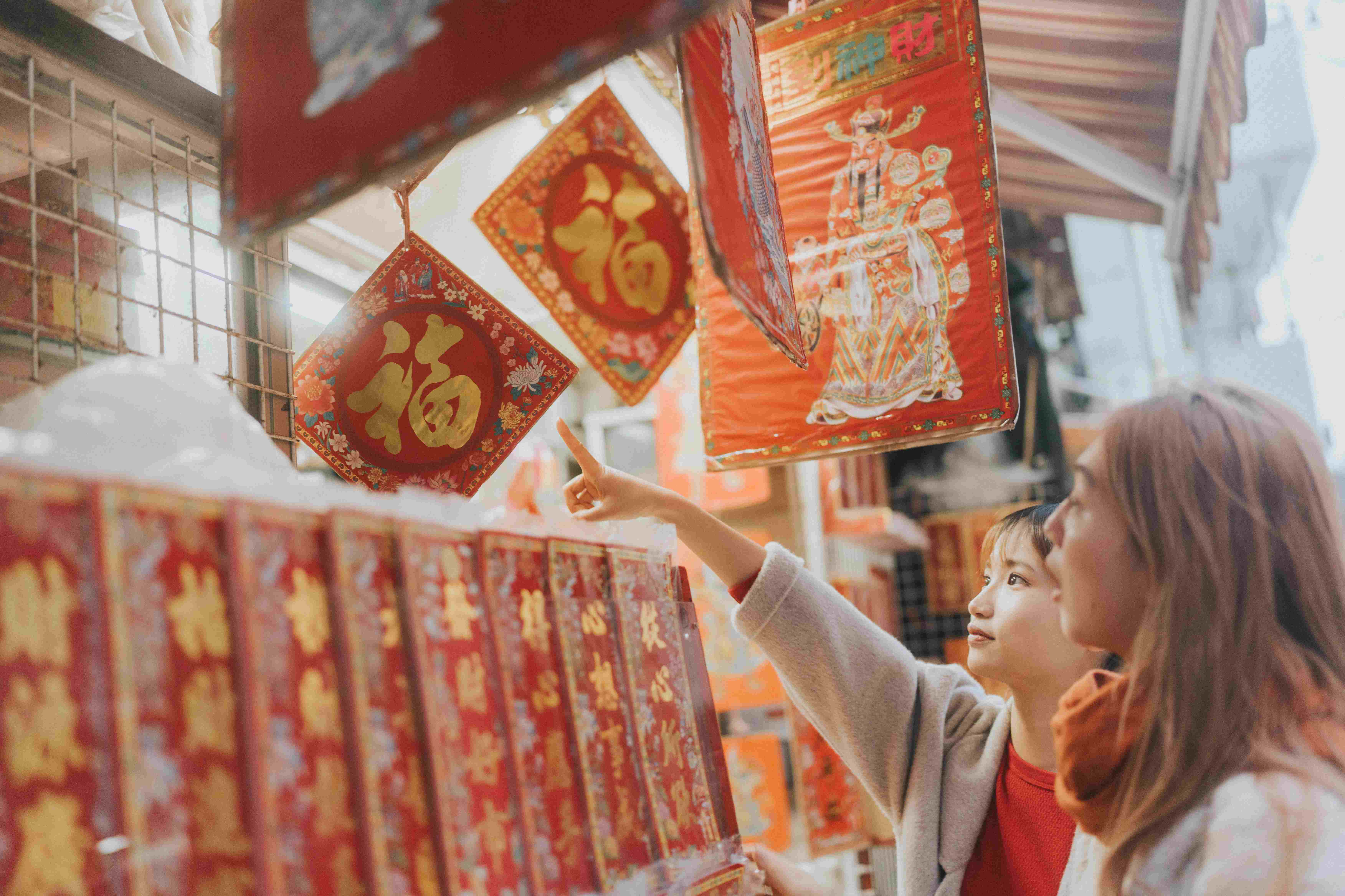 Two women admire decorative red and gold items hanging at a market, pointing at one while browsing the scene.