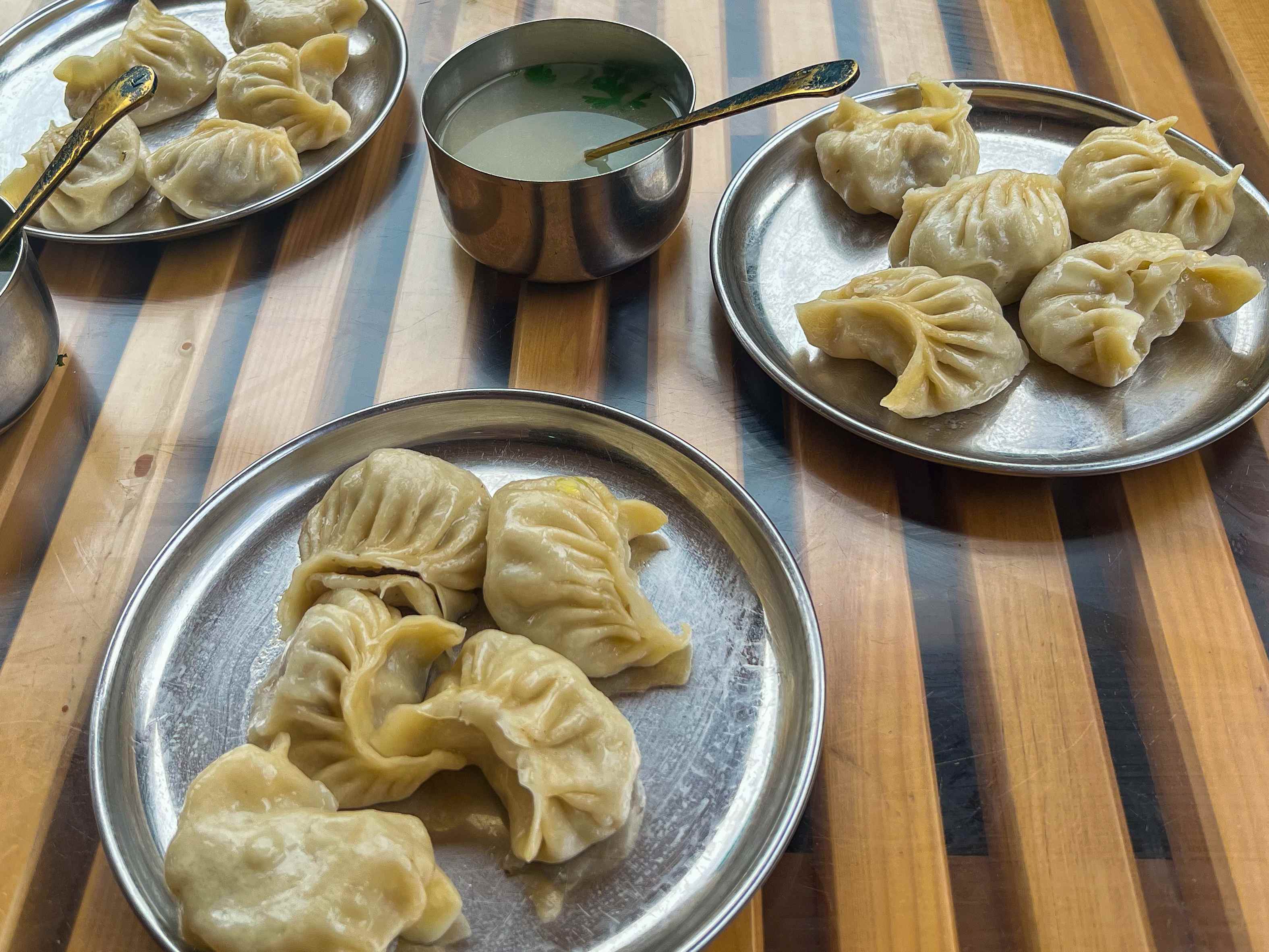 Three metal plates hold steamed dumplings, with a bowl of sauce on a wooden table.