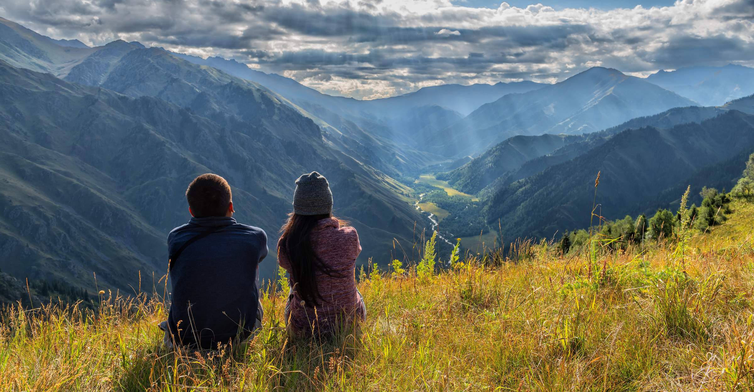 Two people sit on a grassy hillside, gazing at a panoramic view of mountains and valleys under a cloudy sky.