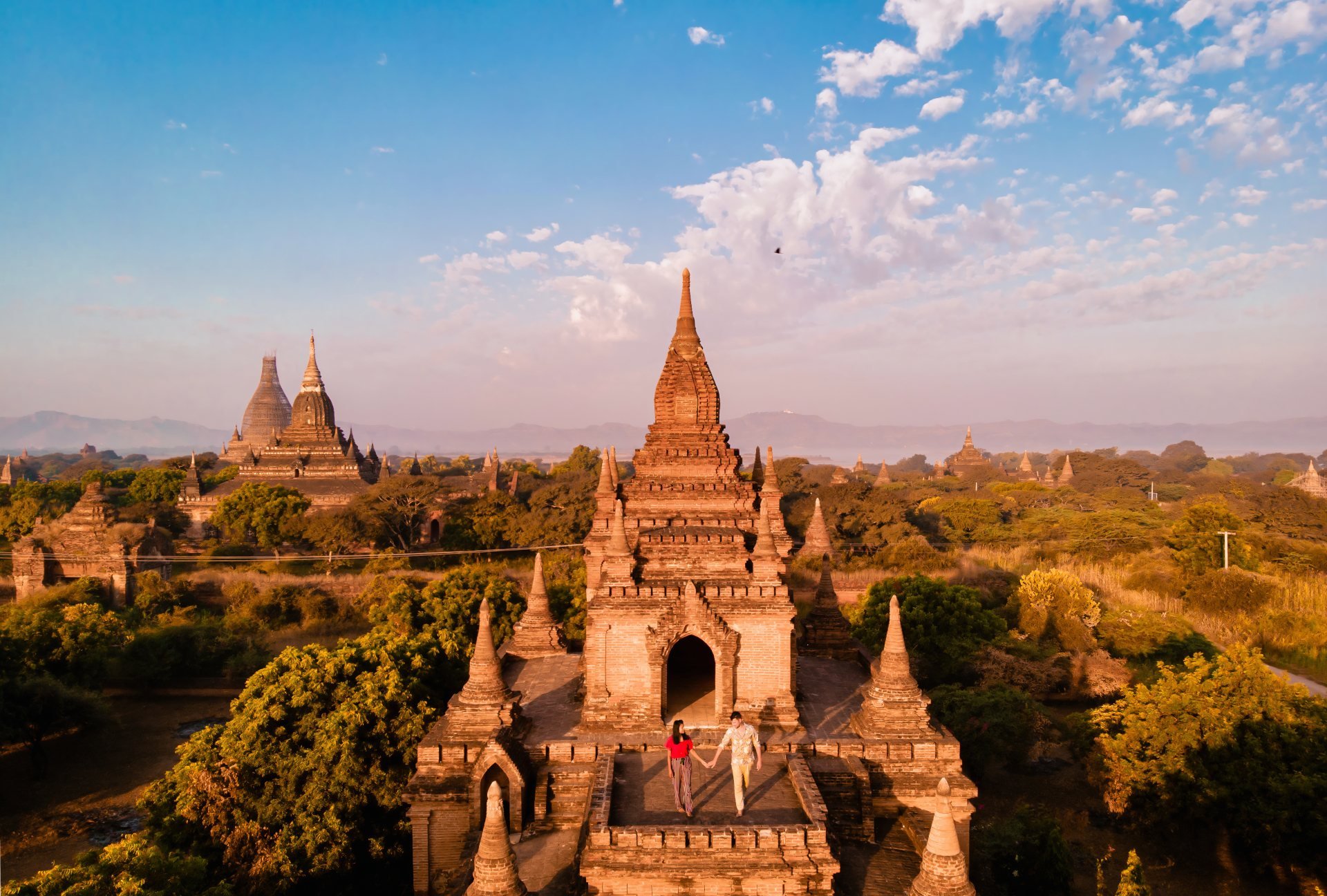 Aerial view of ancient temples in Bagan, Myanmar, with two people exploring among greenery under a blue sky.