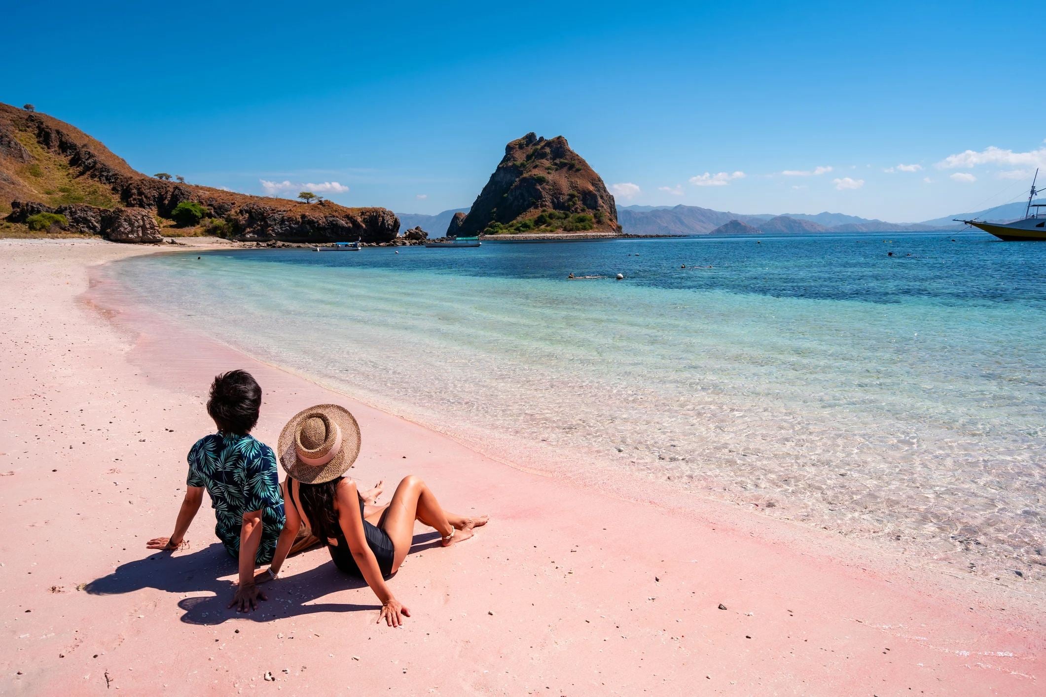 Two people relax on a pink beach, looking out at clear blue water and a rocky island in the distance under a sunny sky.