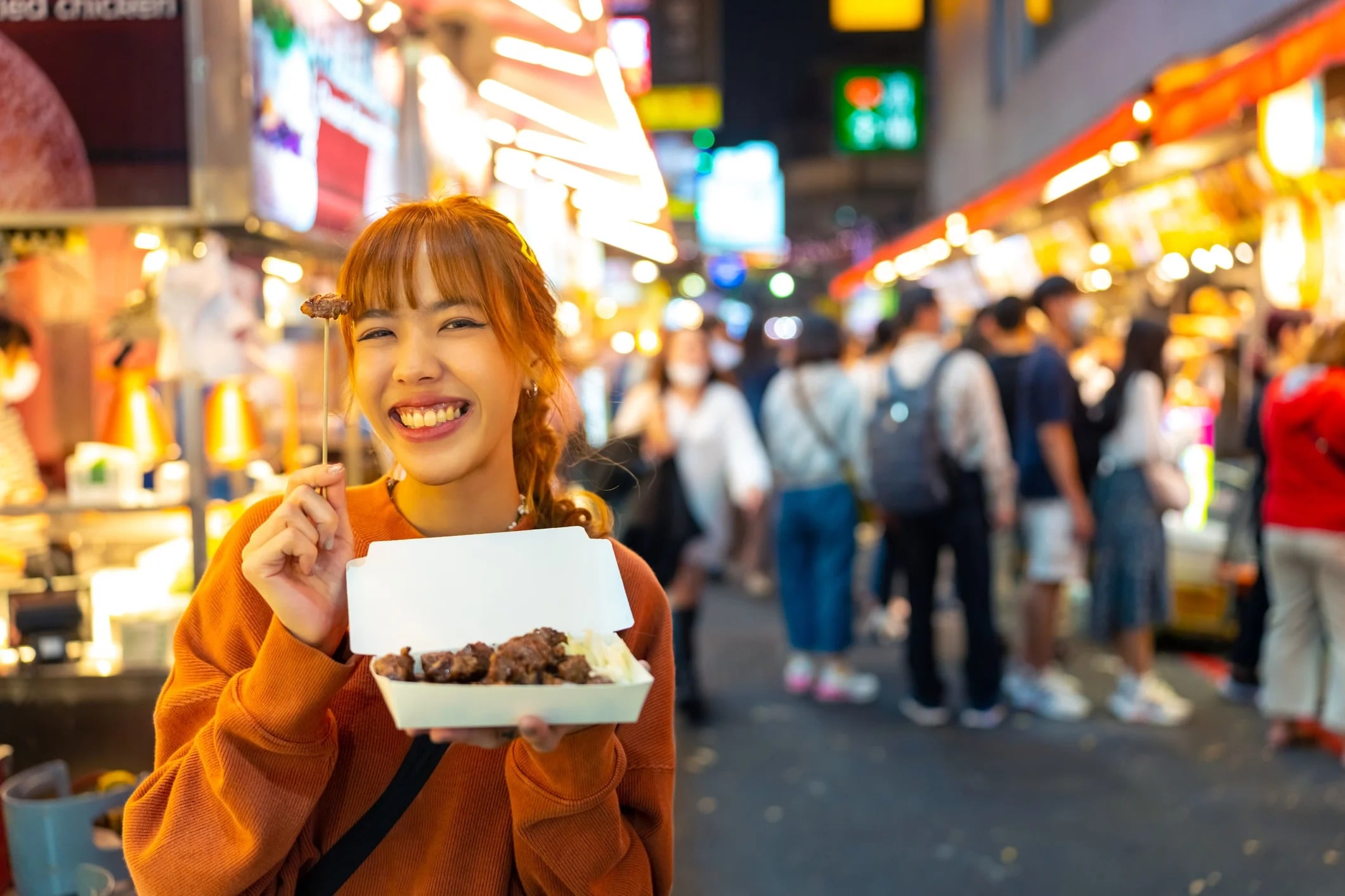 A smiling woman holds a takeout box of food at a busy night market filled with colorful stalls and crowds.
